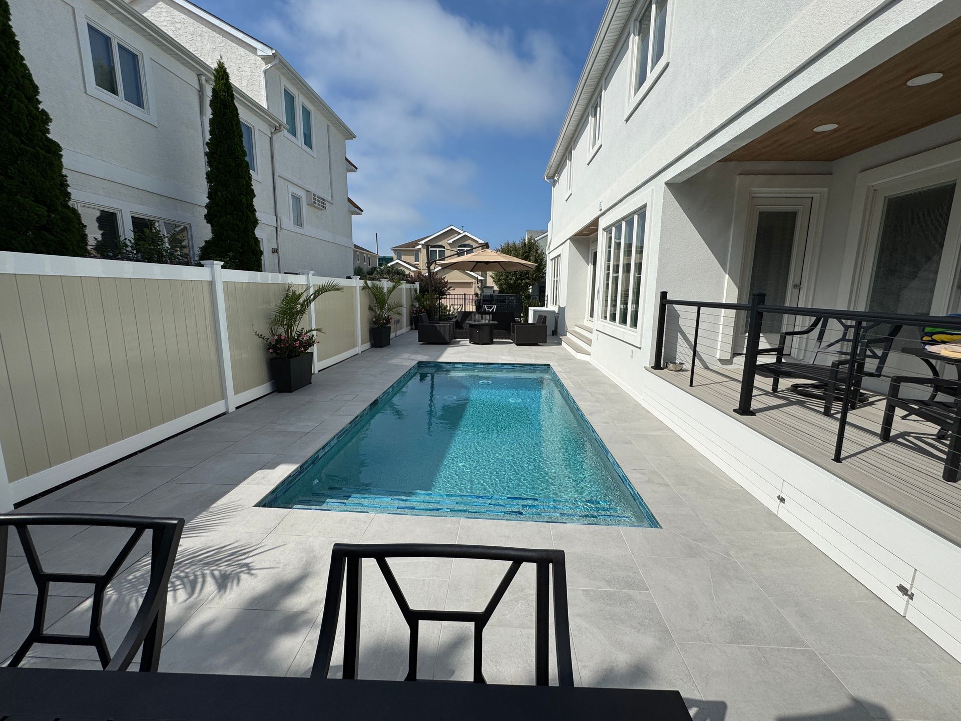Narrow backyard pool between white buildings, with patio chairs and clear blue water.