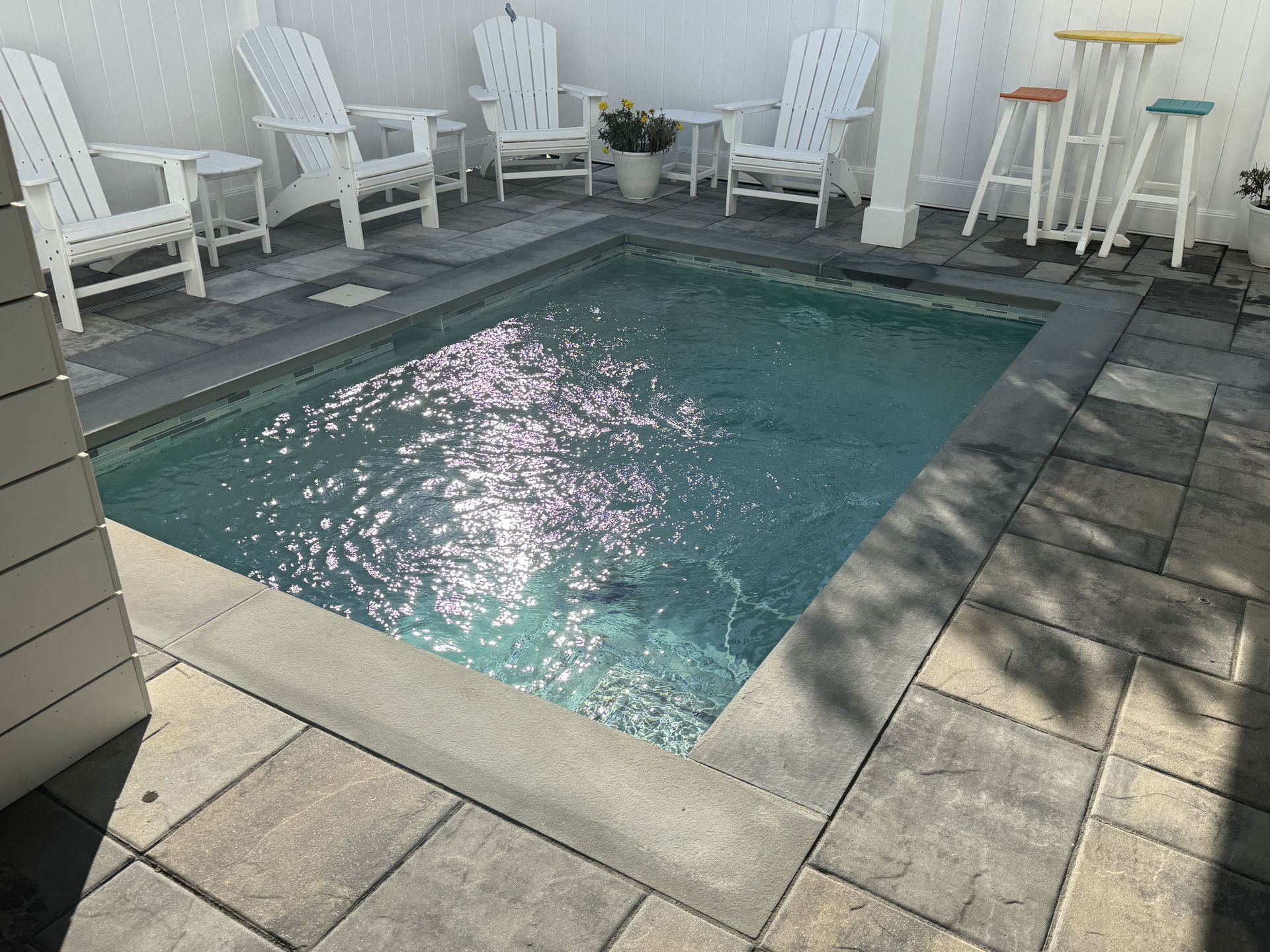 Small backyard pool with blue water, white lounge chairs, and stools on a stone patio.