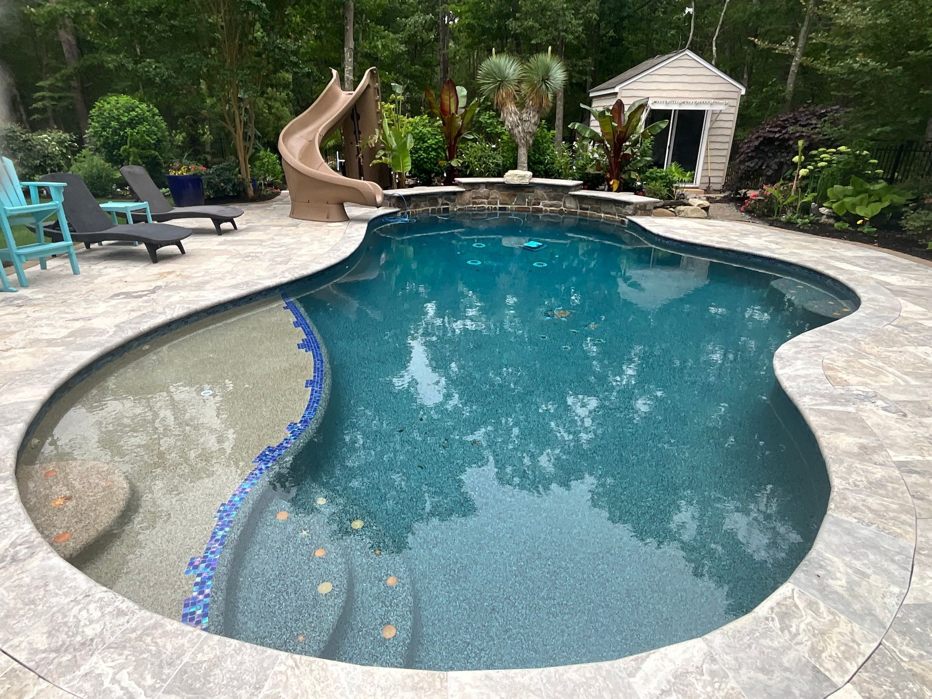 Curved backyard swimming pool with stone deck, slide, lounge chairs, and lush trees nearby