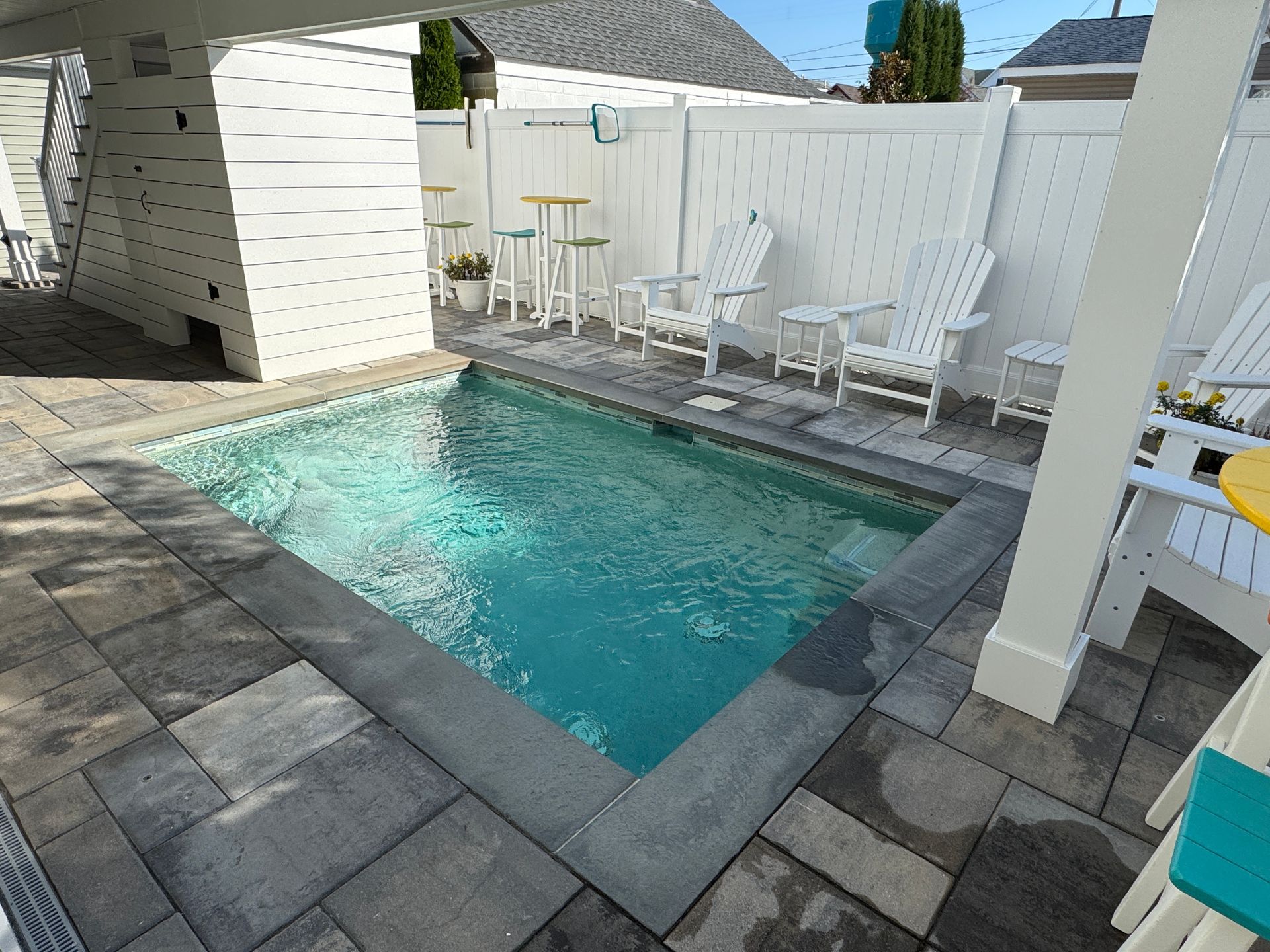 Small backyard pool with gray tile decking and white lounge chairs beside a white fence