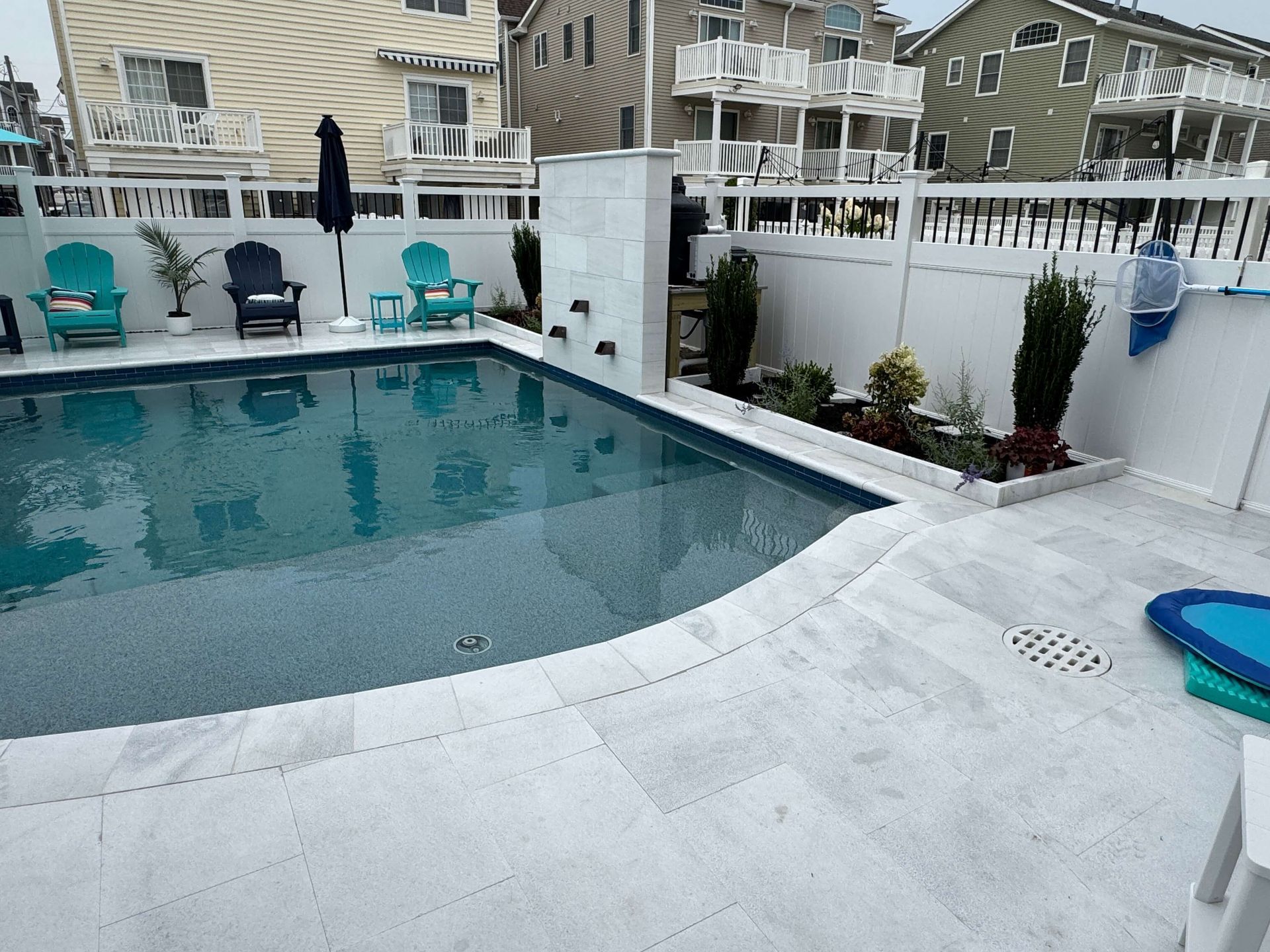 Backyard swimming pool with white fence, patio chairs, and townhouses in the background