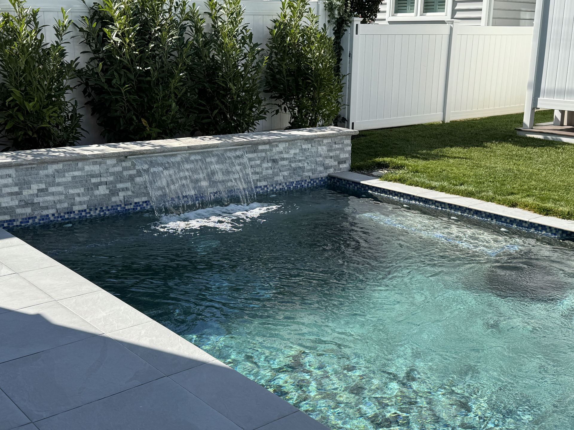 Modern backyard pool with turquoise water, stone coping, white fence, and green lawn.
