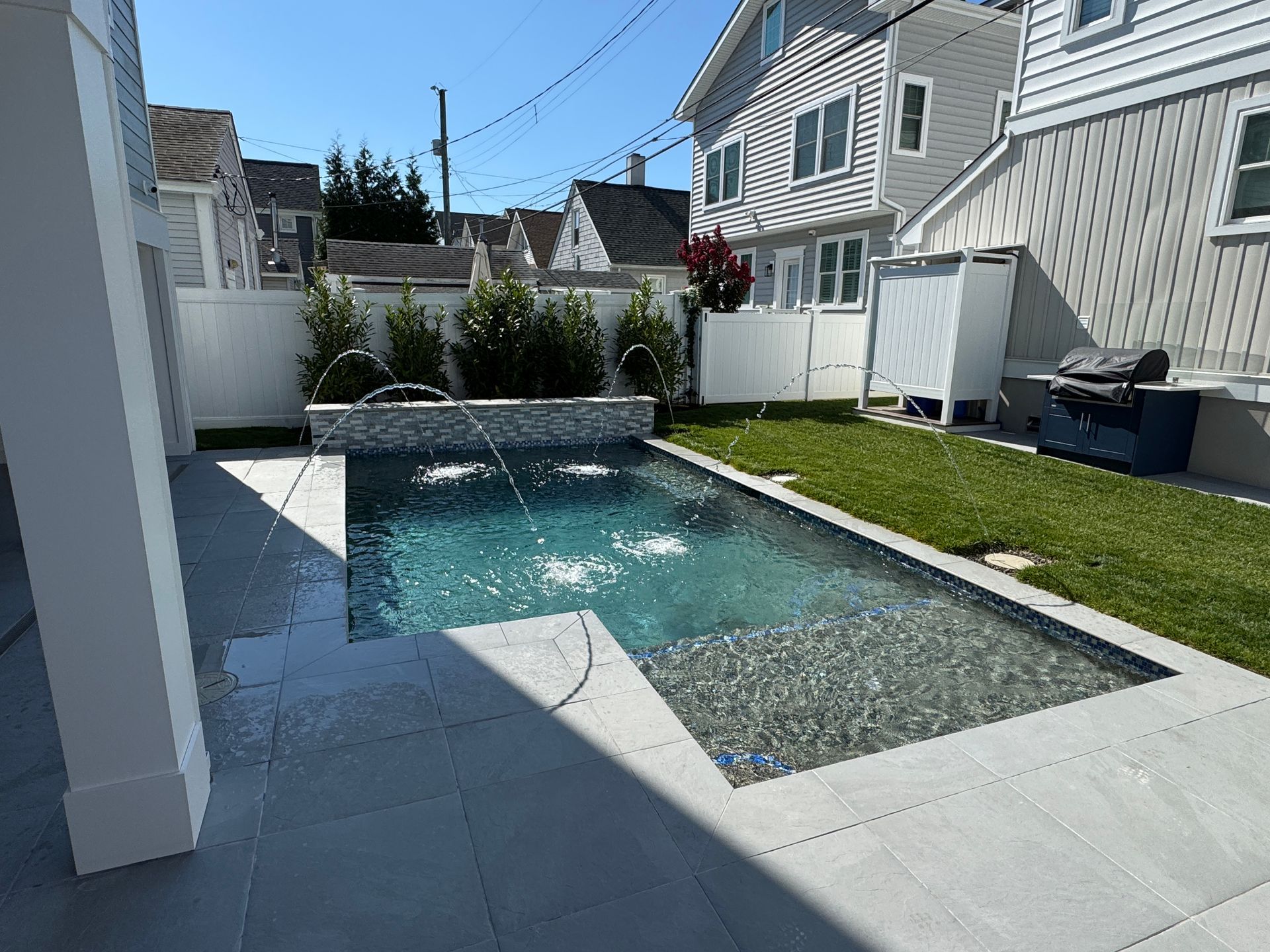 Narrow backyard with a small rectangular pool, gray patio tiles, and white house siding under a blue sky