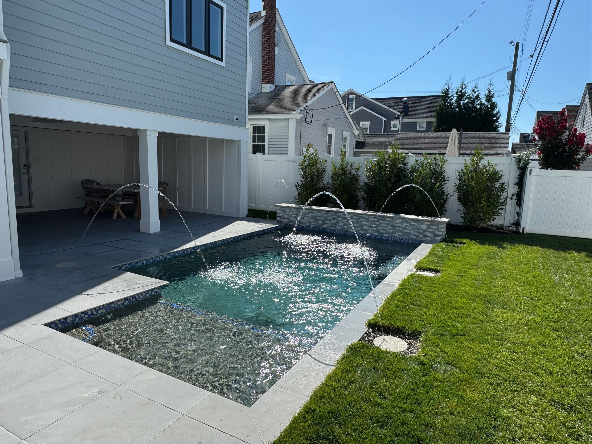 Backyard patio with a small rectangular fountain pool beside a green lawn and white fence
