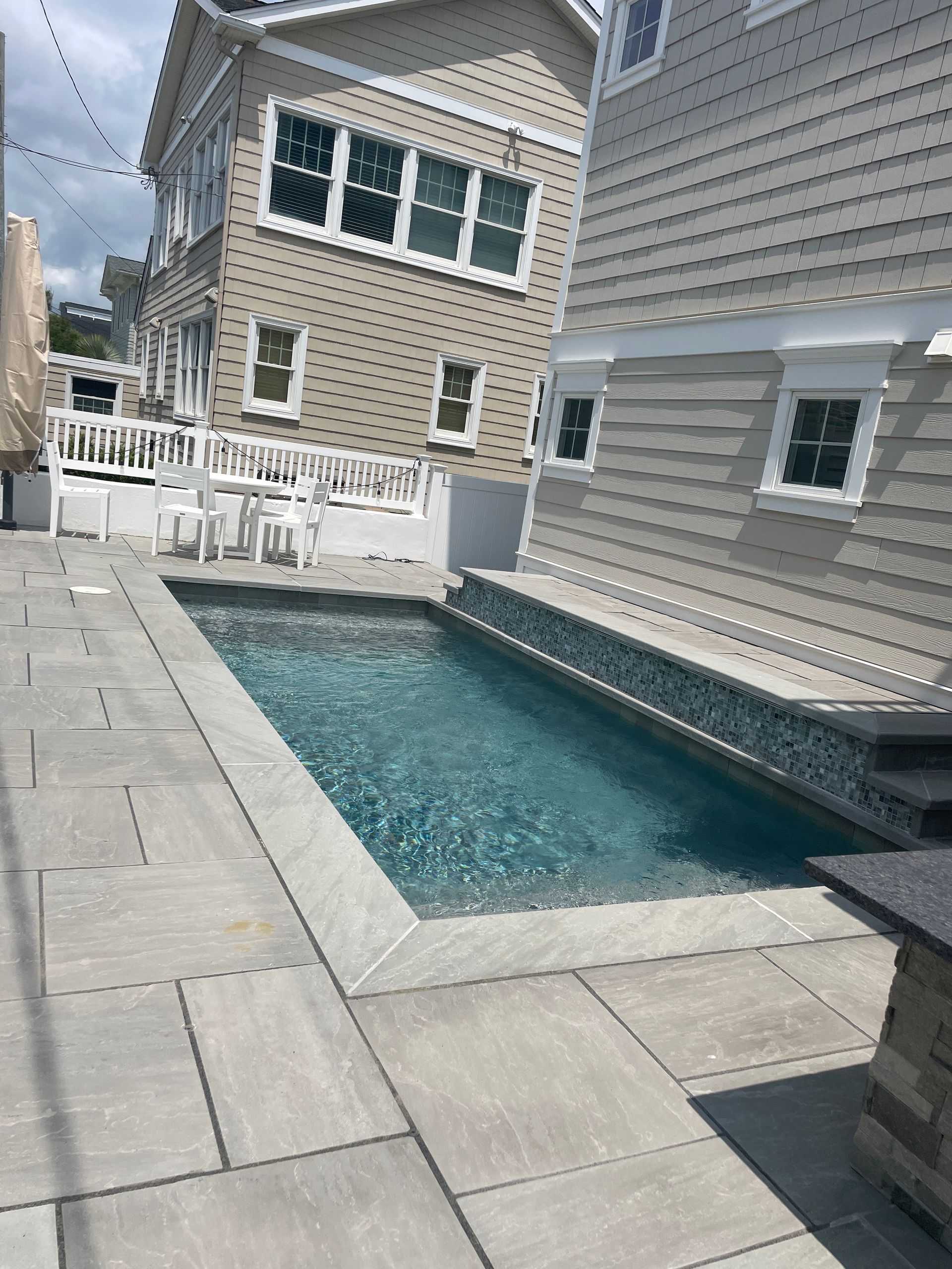 Narrow backyard with a small rectangular pool, gray stone patio, and neighboring houses.