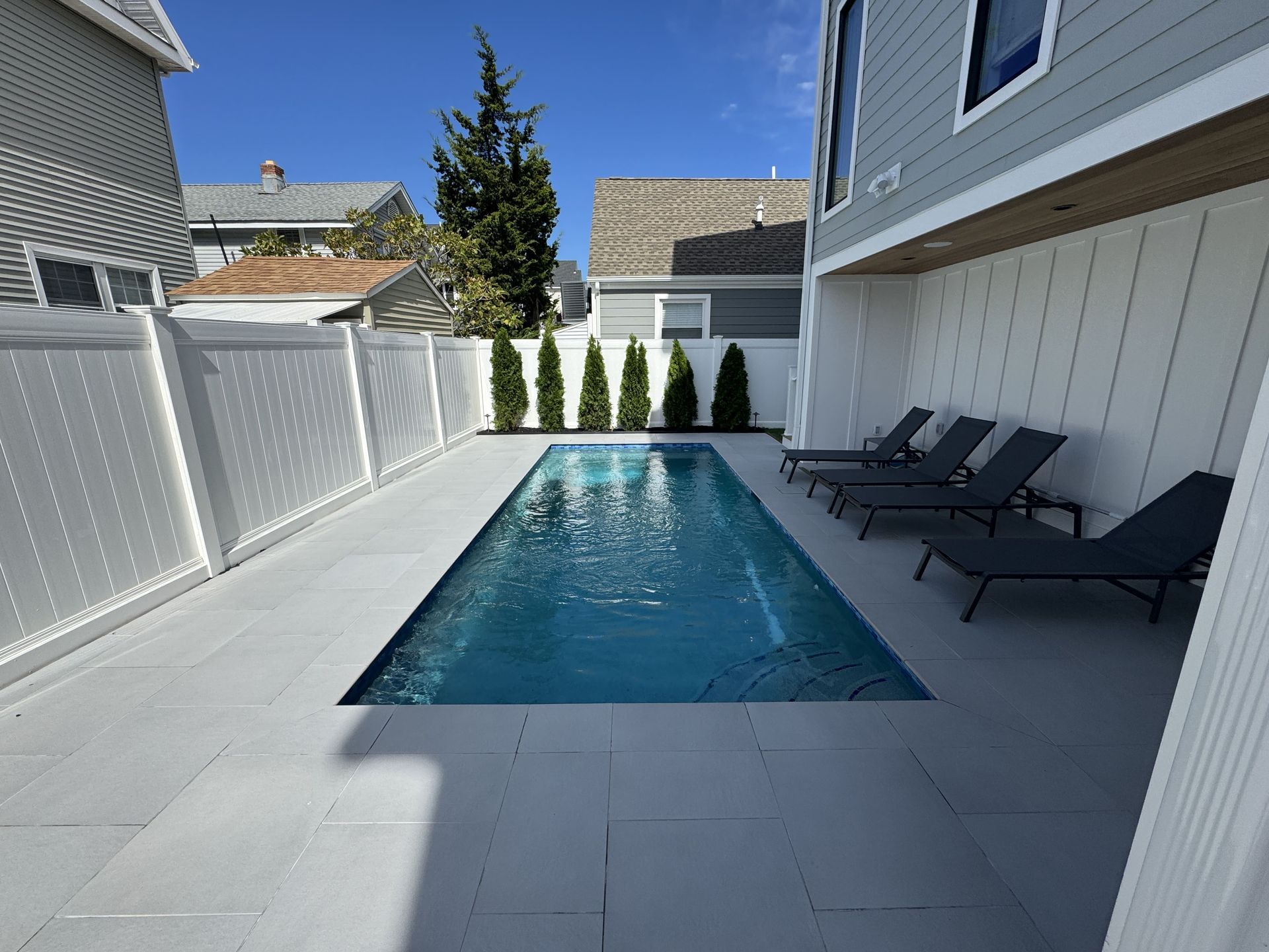 Narrow backyard with a rectangular swimming pool, lounge chairs, white fencing, and house siding.