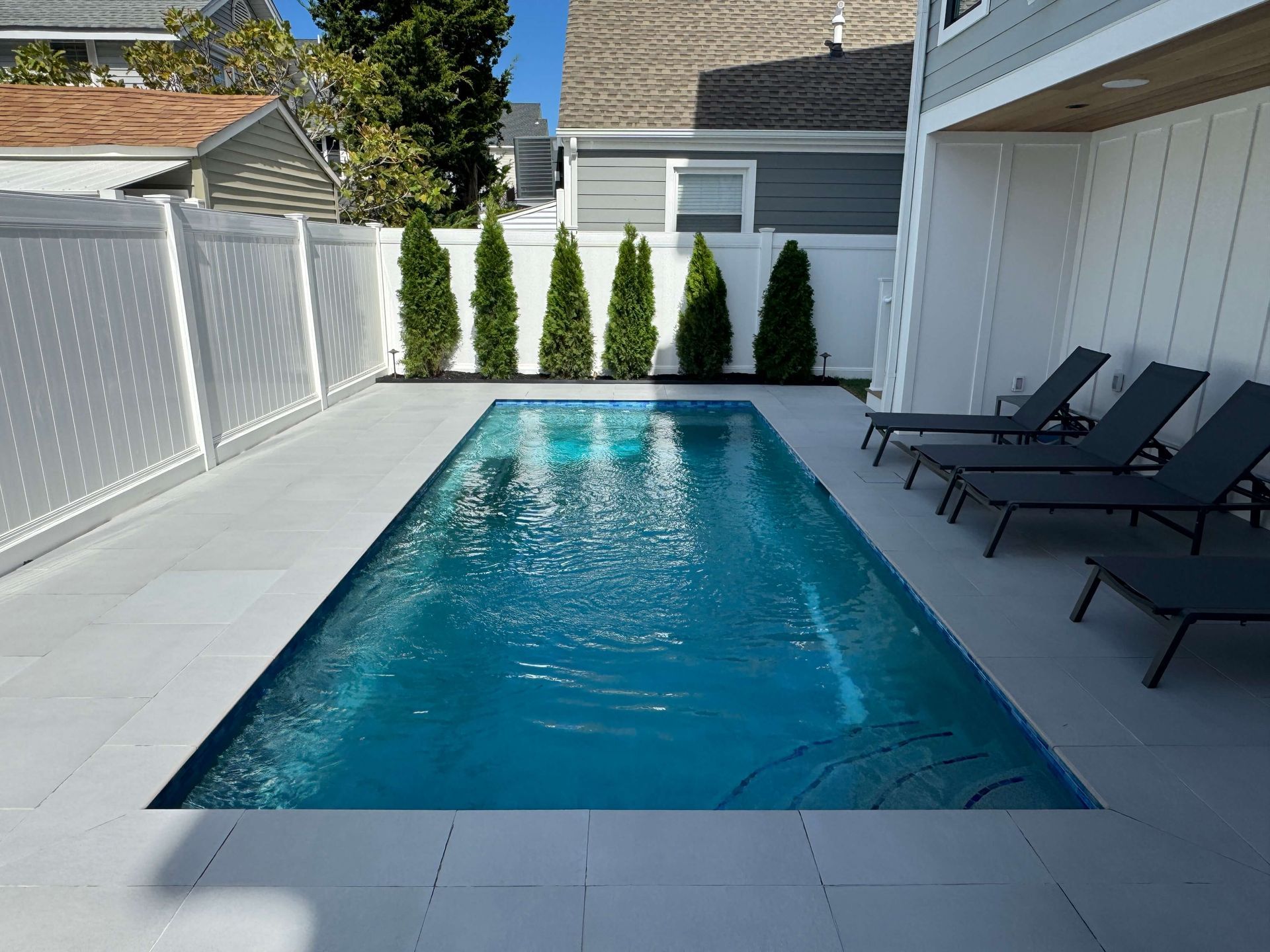 Narrow backyard pool with blue water, white deck, lounge chairs, and tall green shrubs along the fence