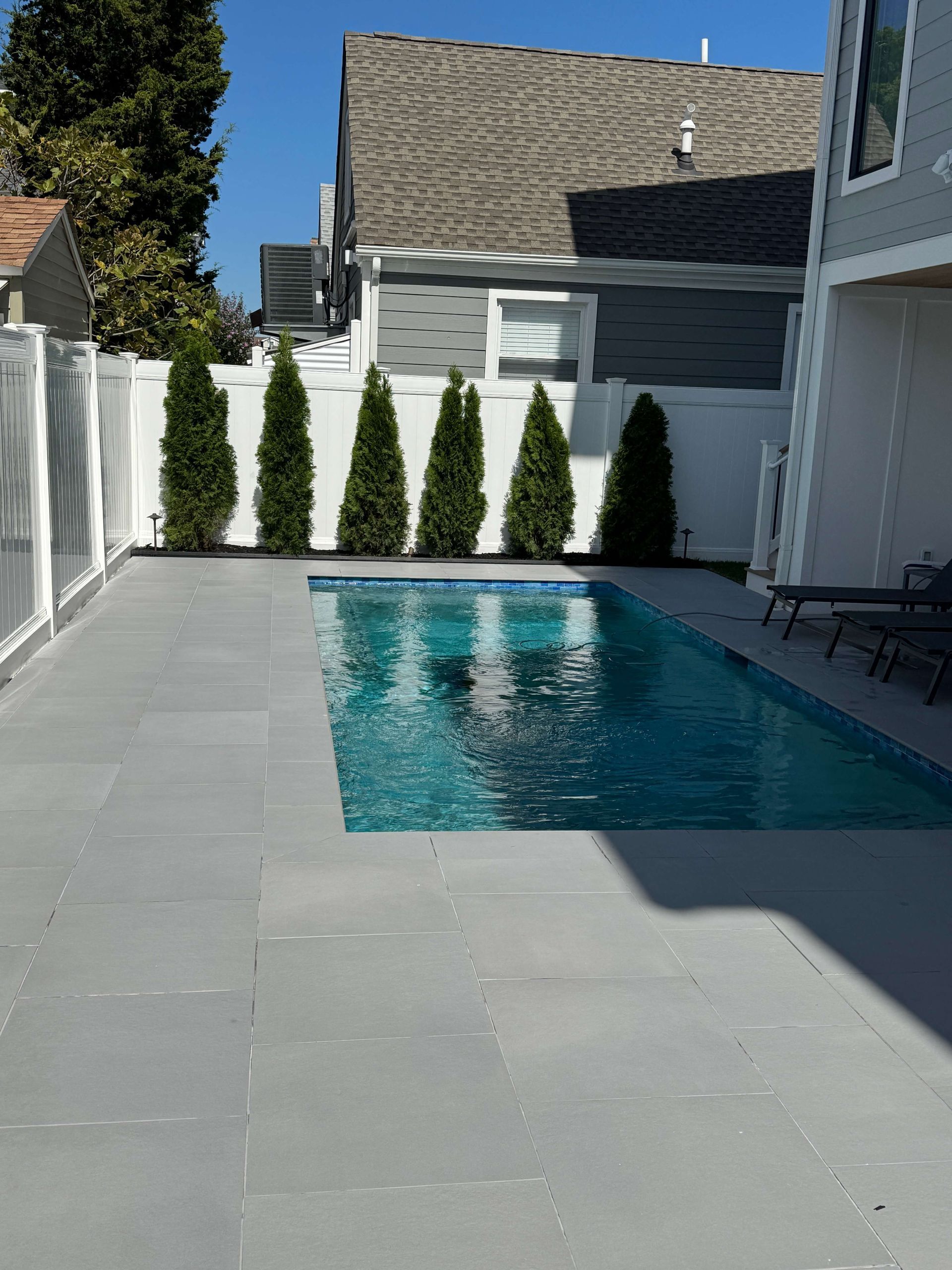 Backyard with a rectangular swimming pool, gray patio, white fence, and tall evergreens under a clear blue sky