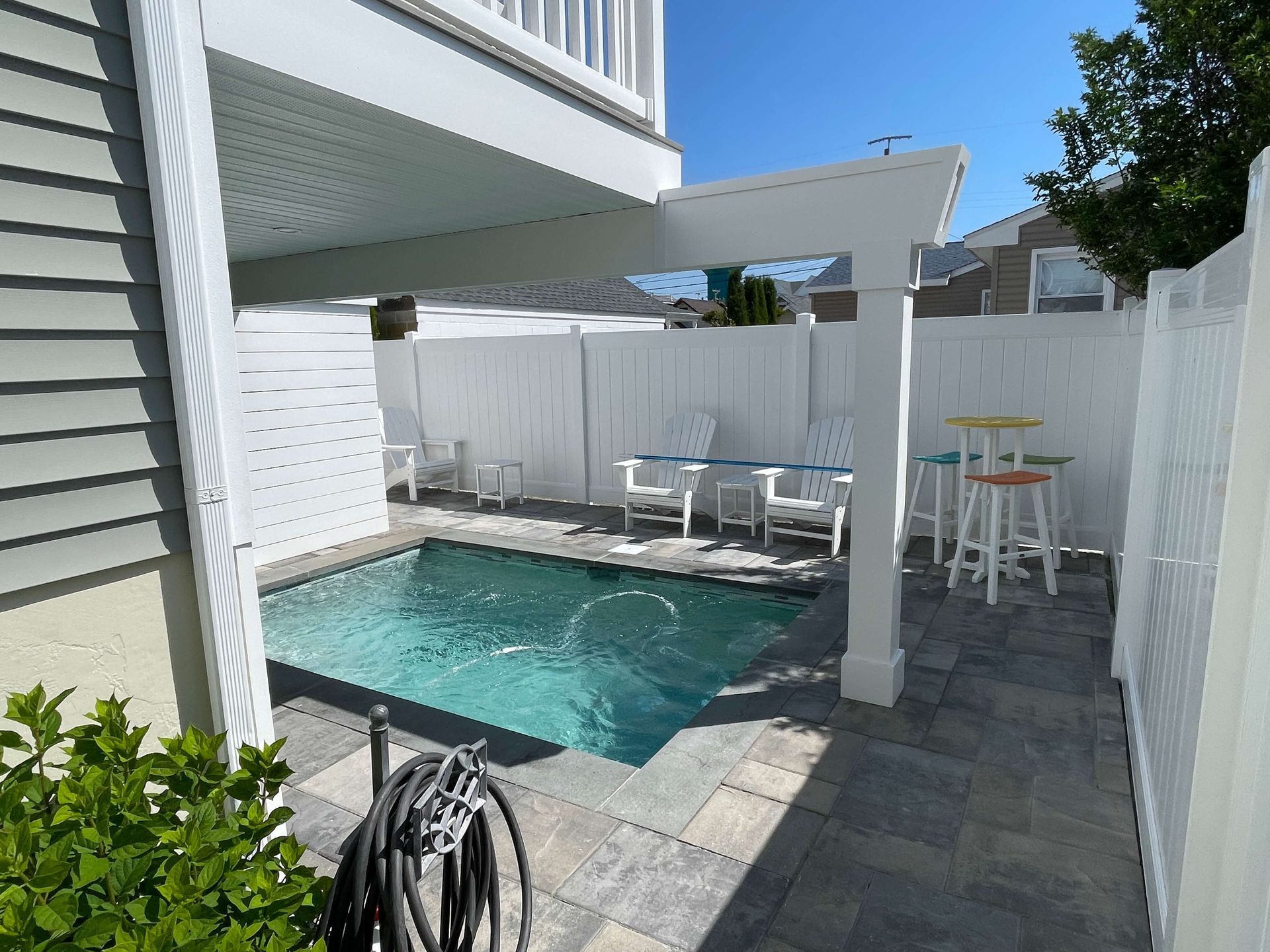 Small private pool with white pergola, patio seating, and gray pavers beside a house.