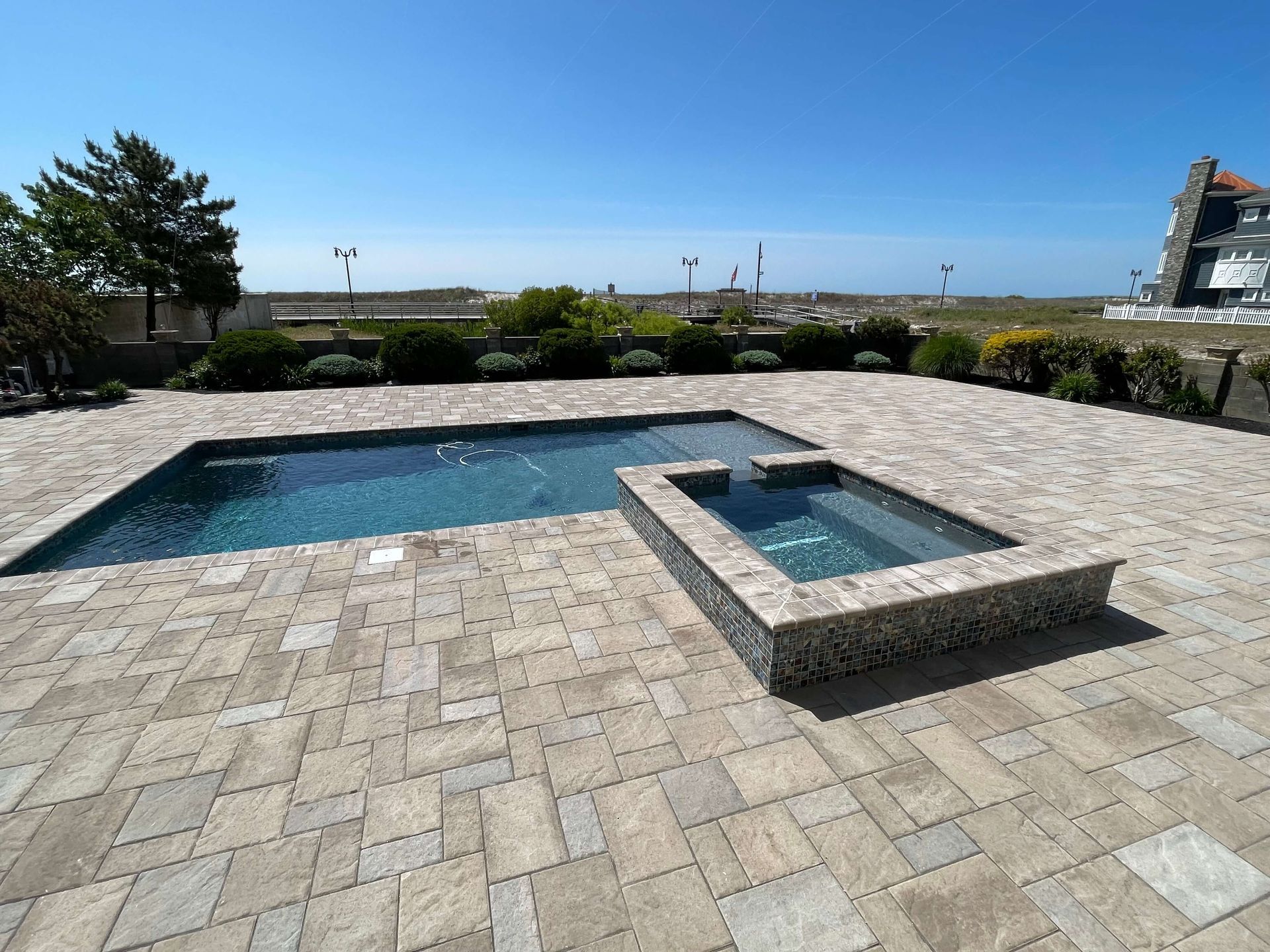Modern backyard pool with stone patio, small hot tub, and ocean view under a clear blue sky