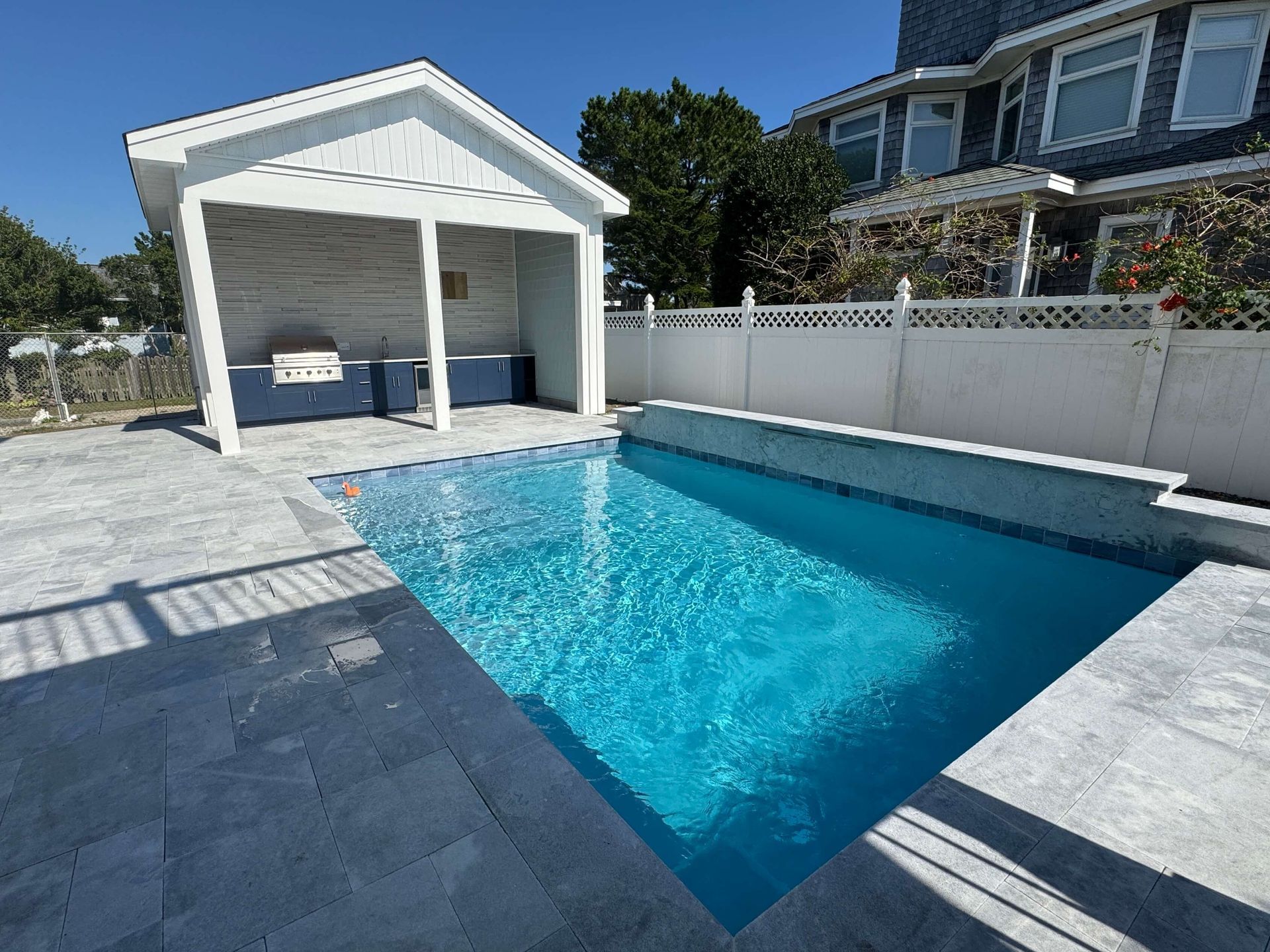 Backyard swimming pool beside a white cabana and fence in bright sunlight