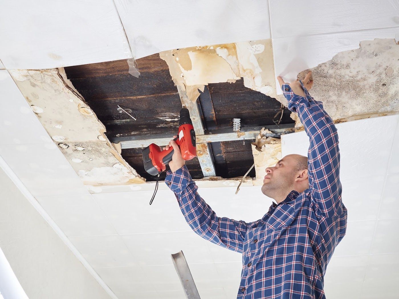 Man repairing water-damaged ceiling with a drill and tools on a ladder