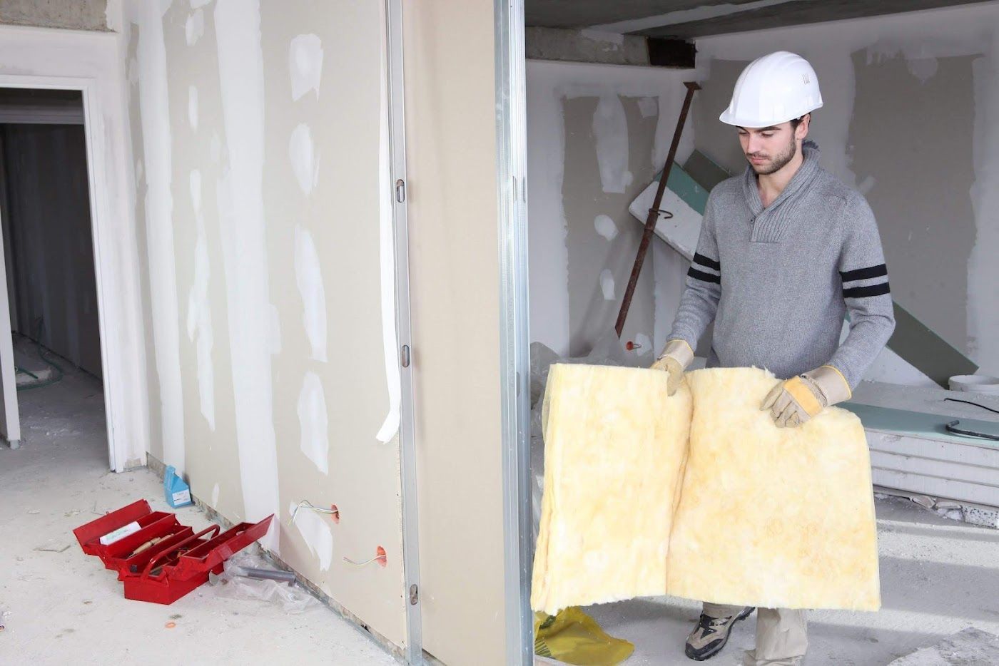 Worker in a hard hat holding insulation boards in an unfinished room with tools on the floor