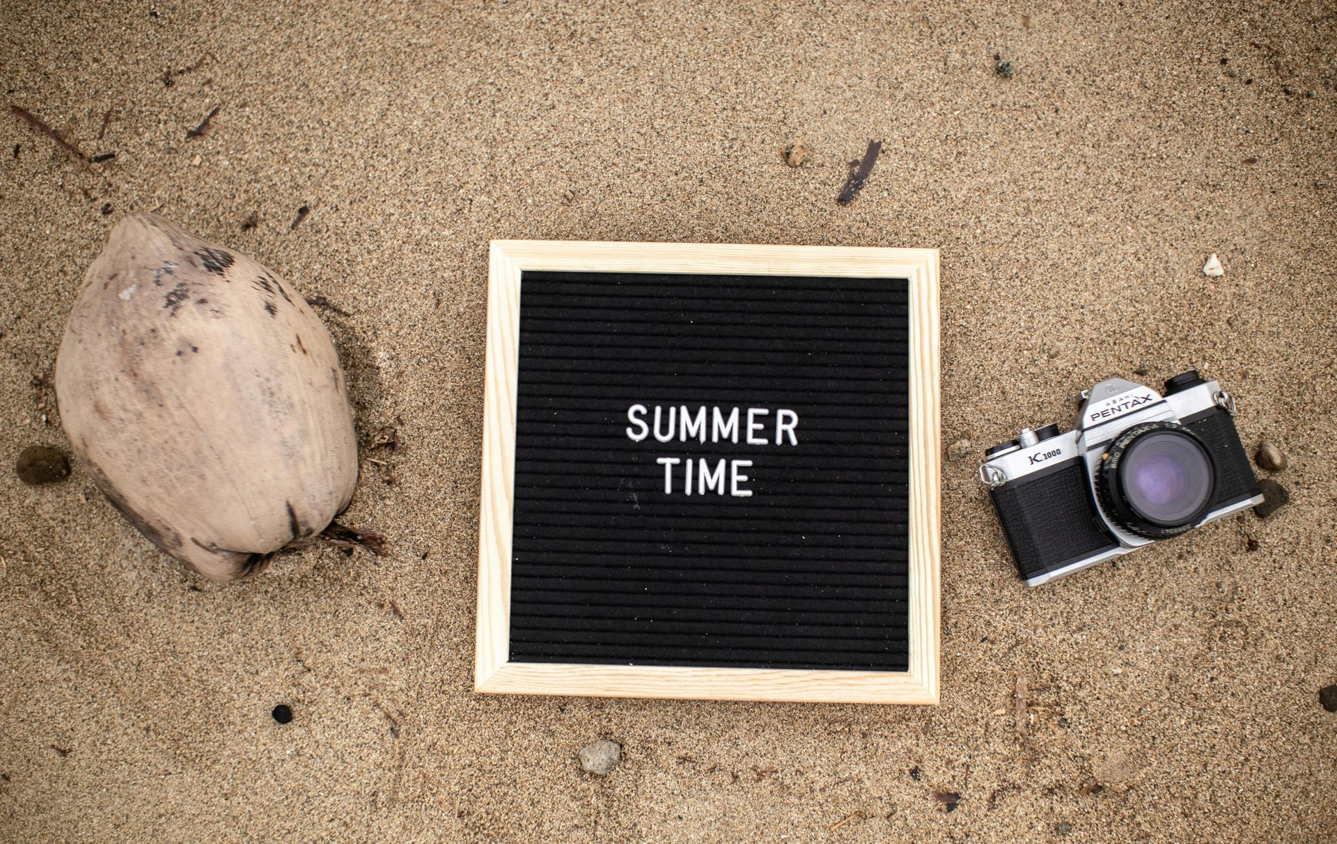 Flat lay on sand with a “SUMMER TIME” sign, a rock, and a vintage camera.