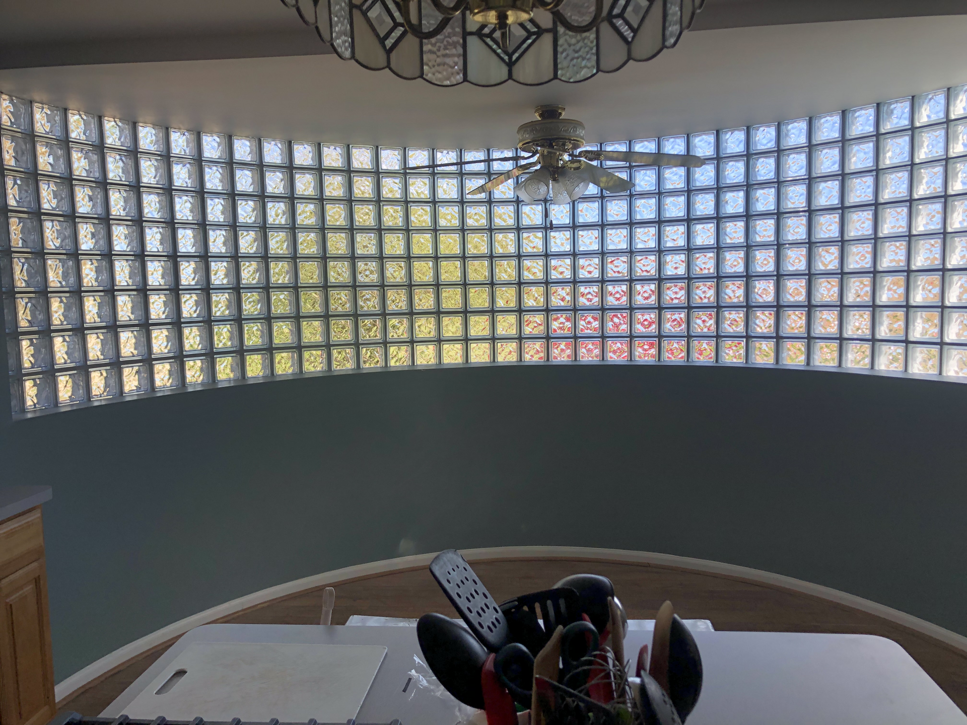 Curved glass block wall over a staircase, with a ceiling fan and chandelier in a bright interior.