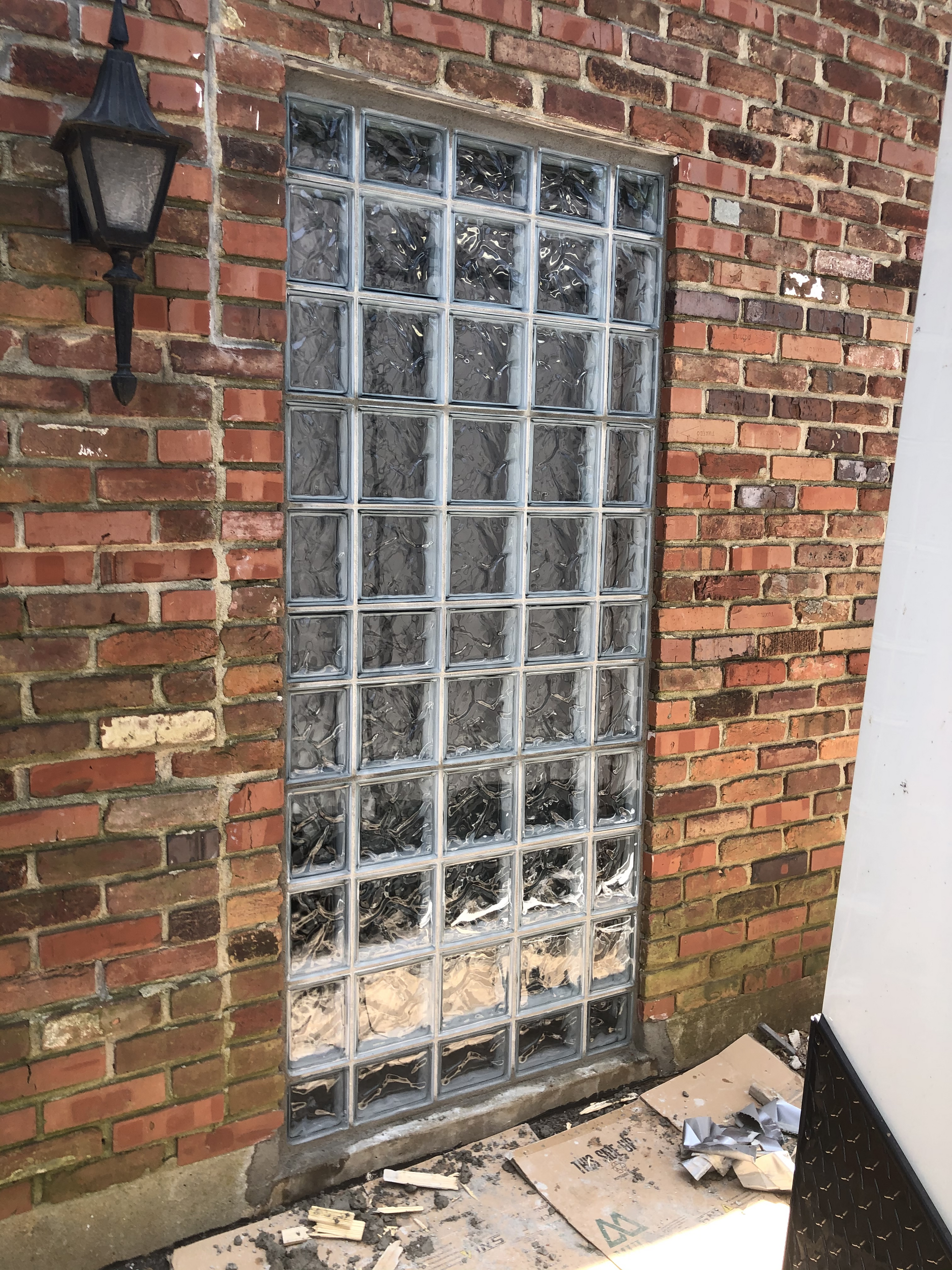 Brick exterior wall with a frosted glass window and black lantern beside a concrete walkway.