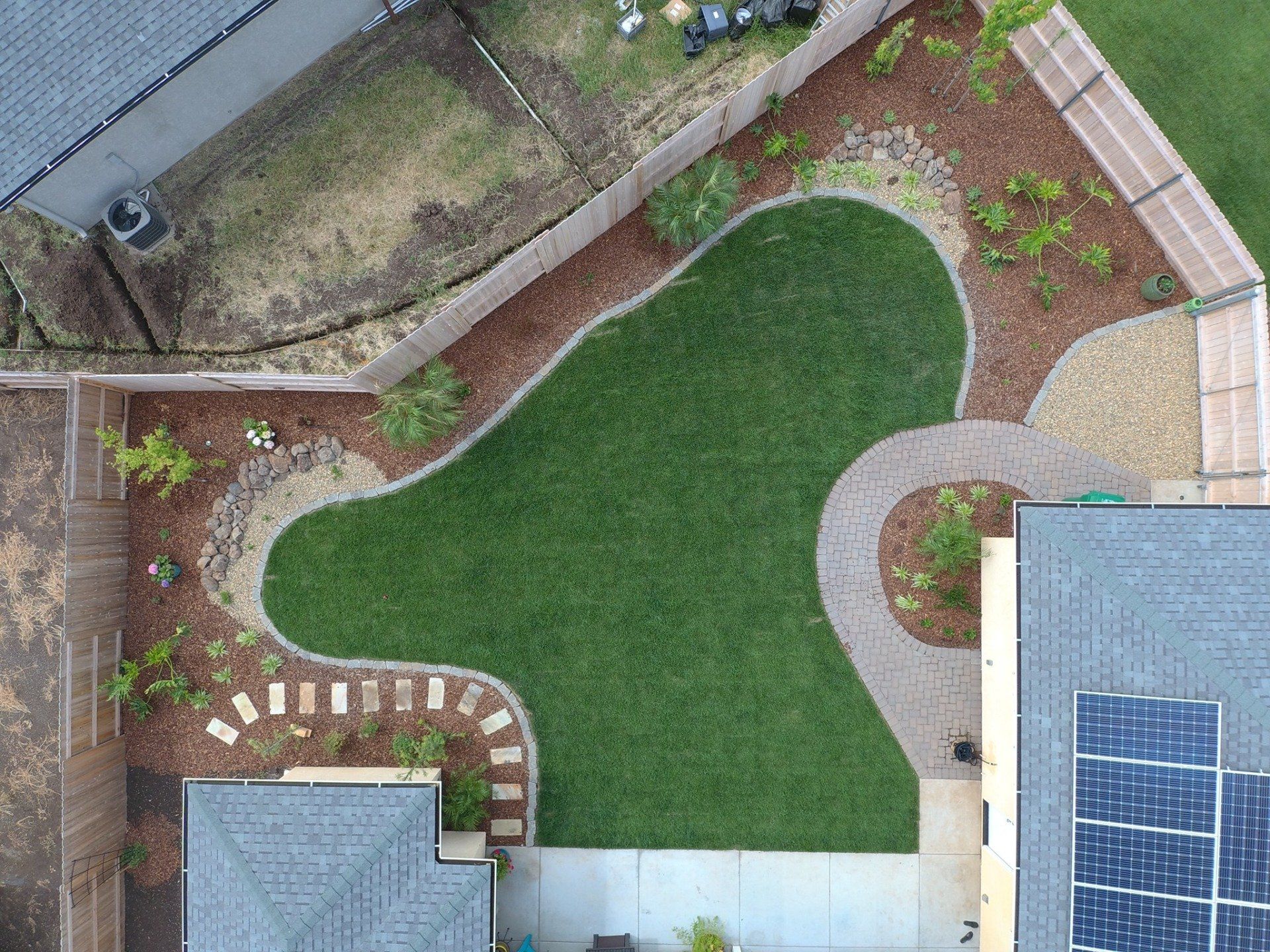 Overhead view of backyard with green lawn, curved stone path, mulch beds with small plants, and solar panels.