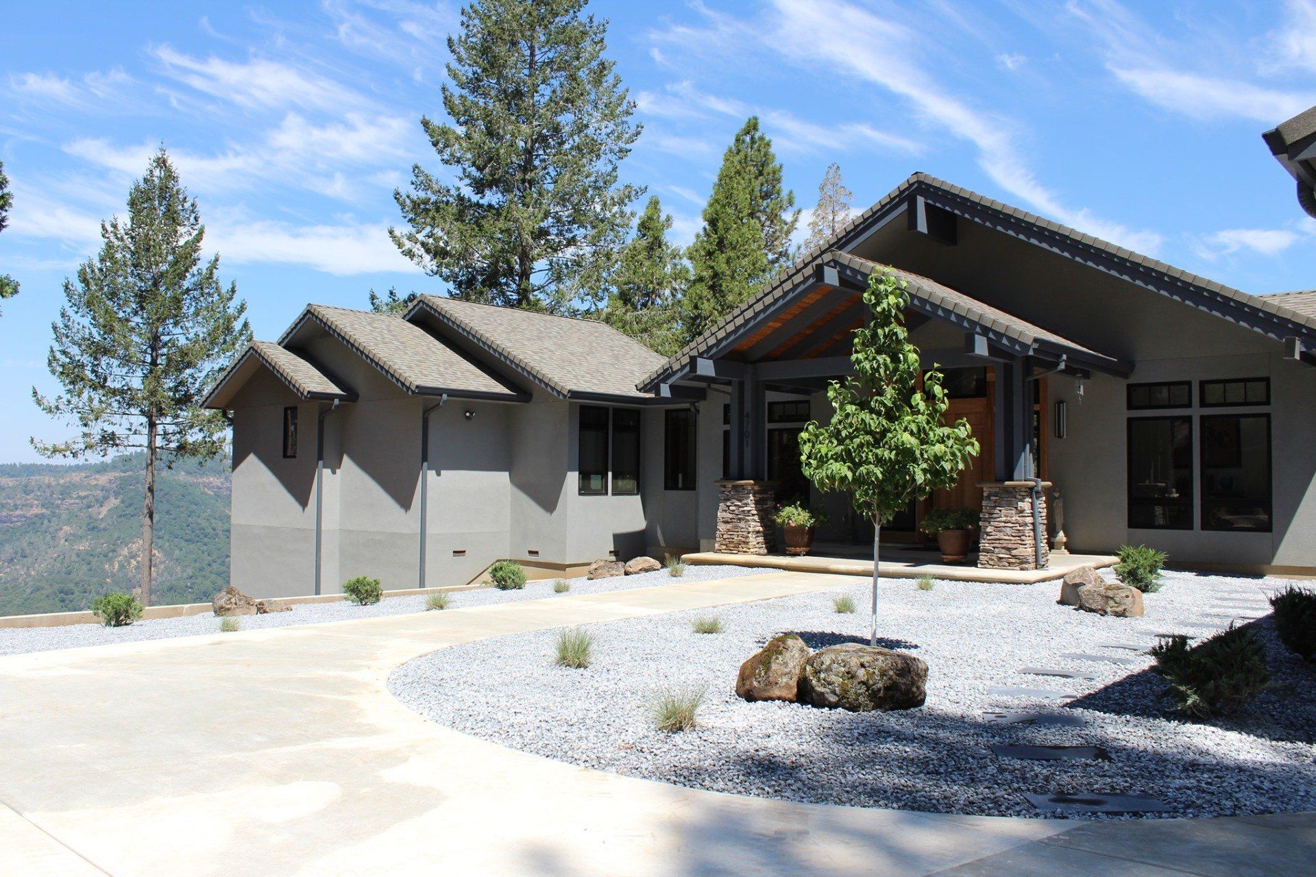 Modern gray house with angled roof, driveway, and landscaping on a hillside under a blue sky.