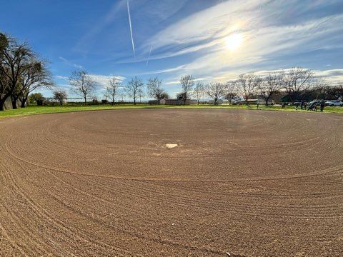Empty dirt baseball field on a sunny day. Trees and lake in the background. Blue sky with clouds.
