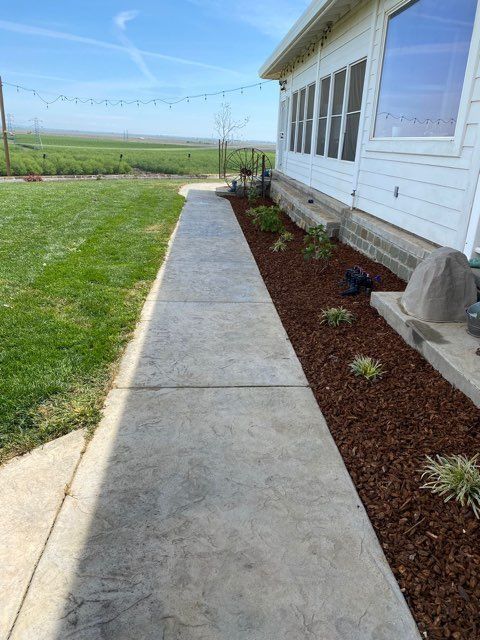 Concrete walkway next to a house with a flower bed and green lawn in a rural setting.