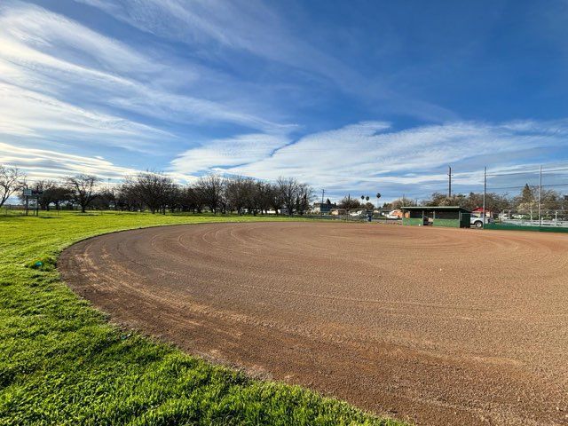 An empty baseball field with a brown dirt infield and green grass under a blue sky.