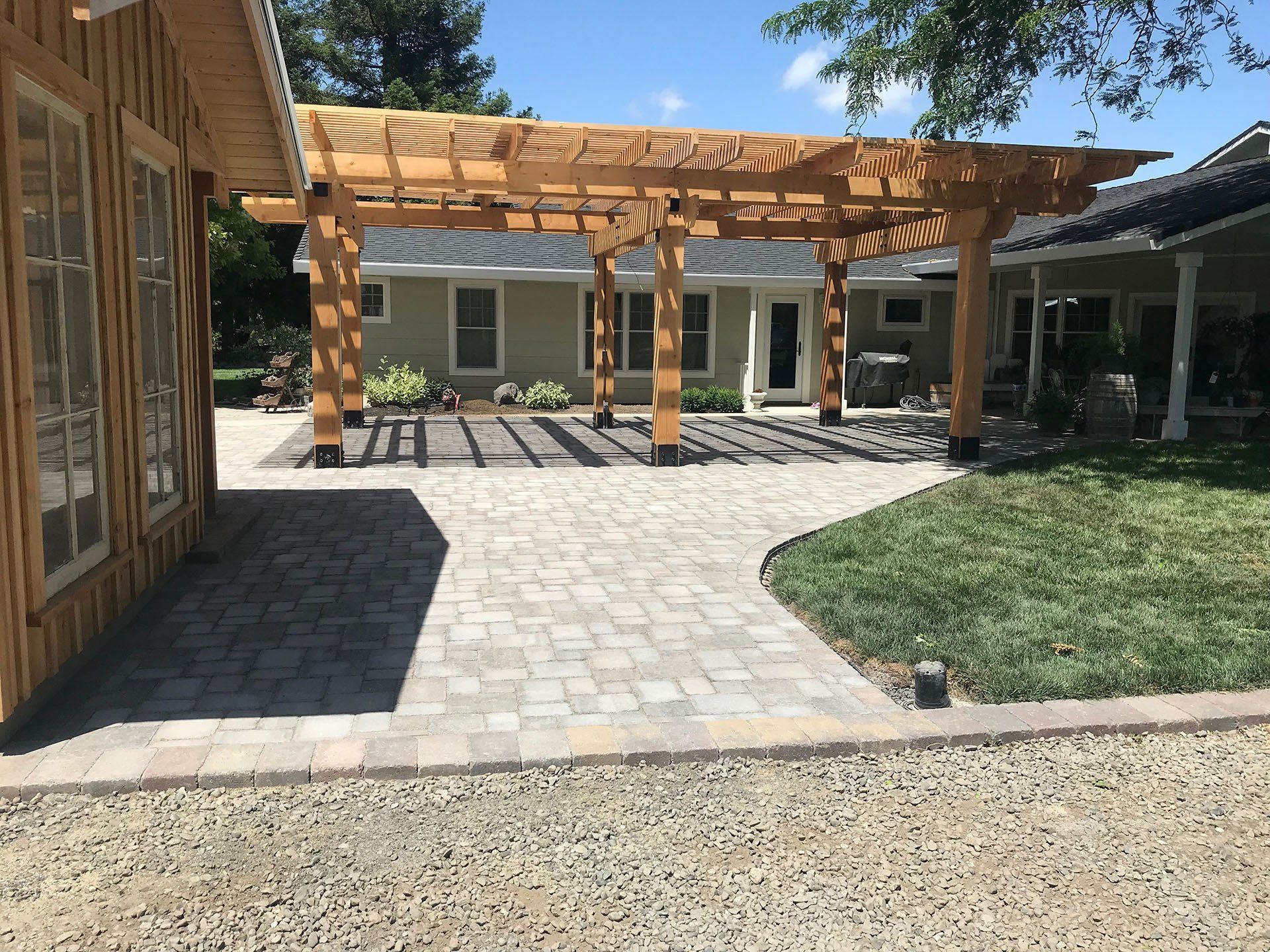 Patio with wood pergola, paved with light-colored stones, next to a house with a grass lawn.