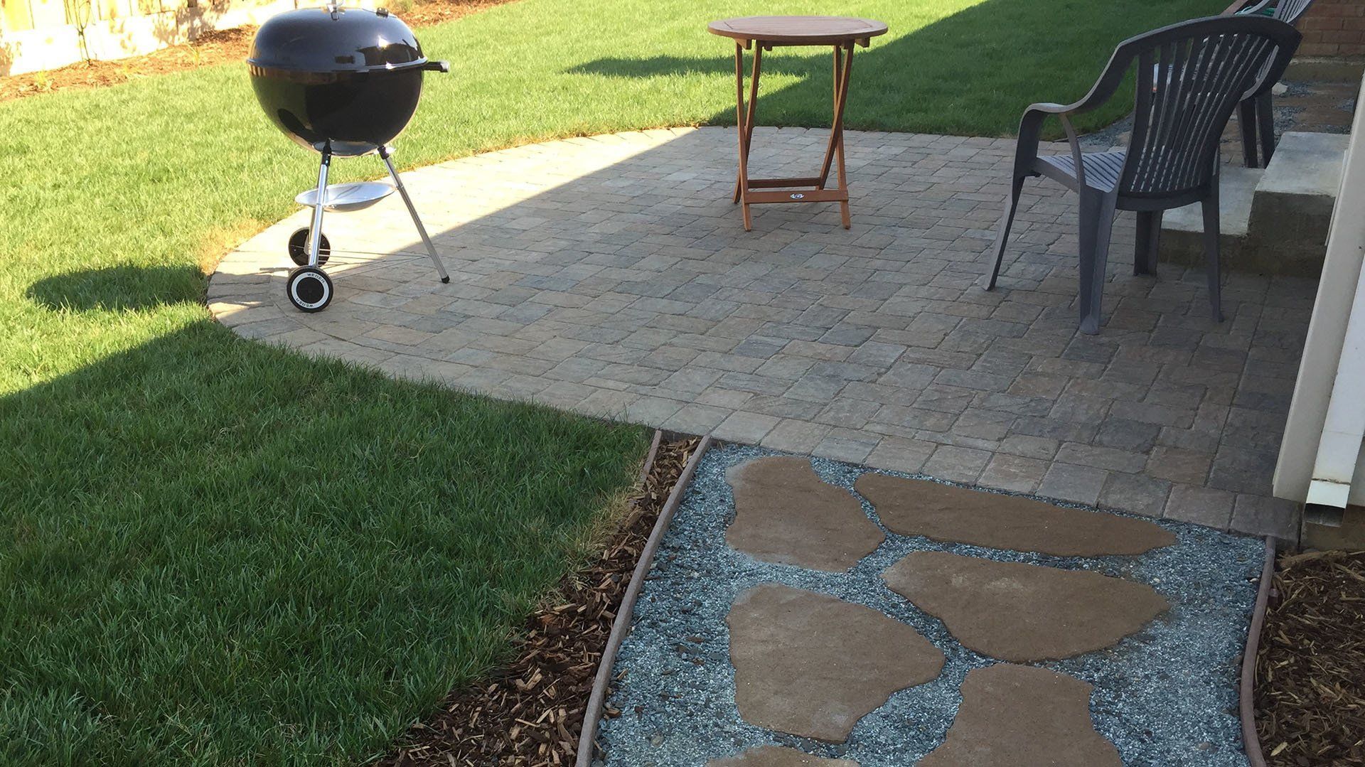 A backyard patio with a grill, table, and chair on pavers, next to grass and a stone path.