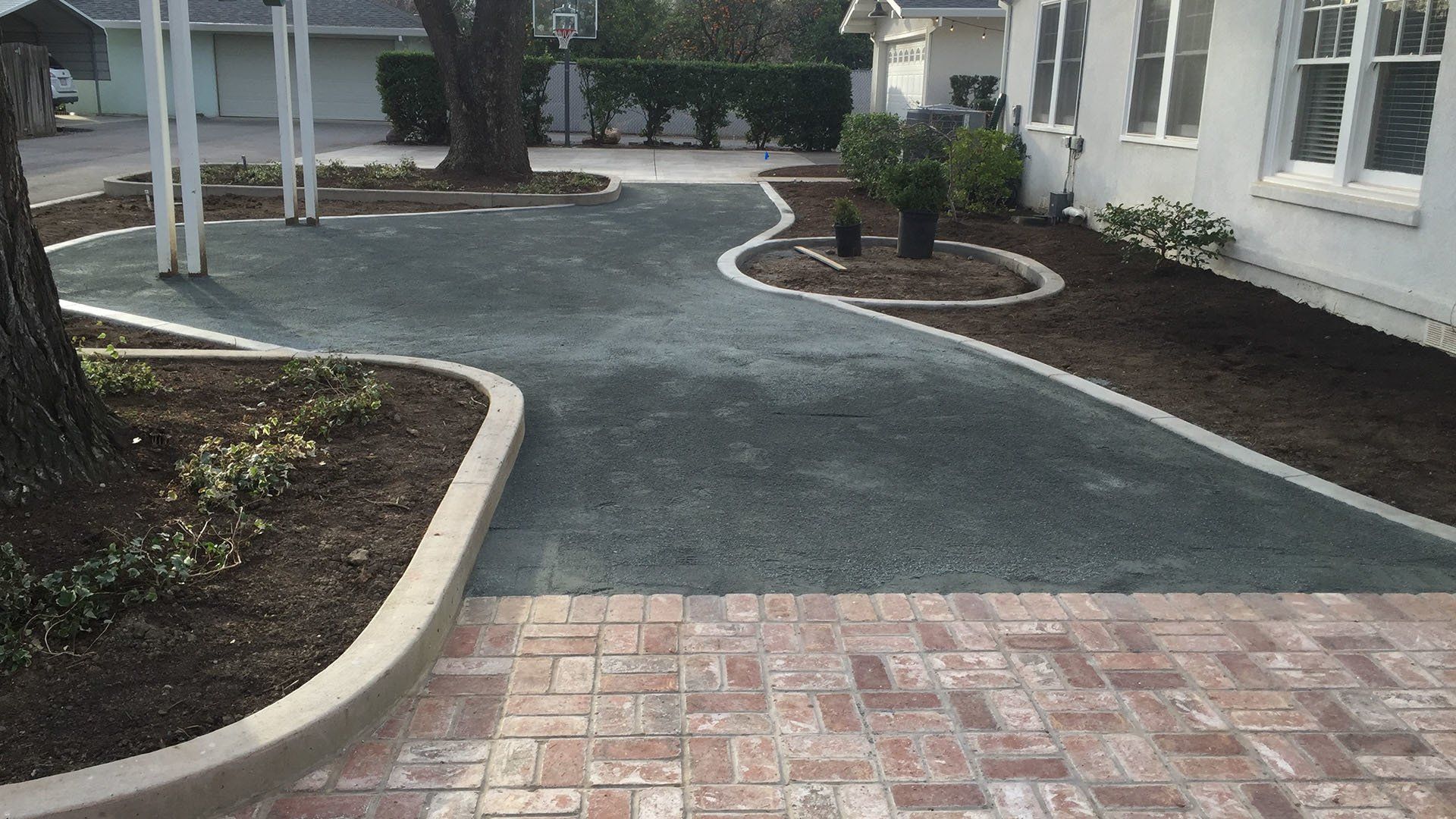 Brick walkway leading to a dark green paved driveway bordered by concrete and mulch beds next to a white house.