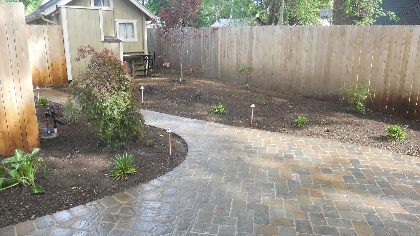 A brick paved pathway curves through a backyard, bordered by fresh mulch, plants, and a wooden fence.