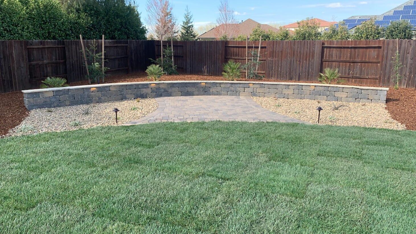 A backyard with a curved stone retaining wall, gravel path, grass lawn, and wooden fence.