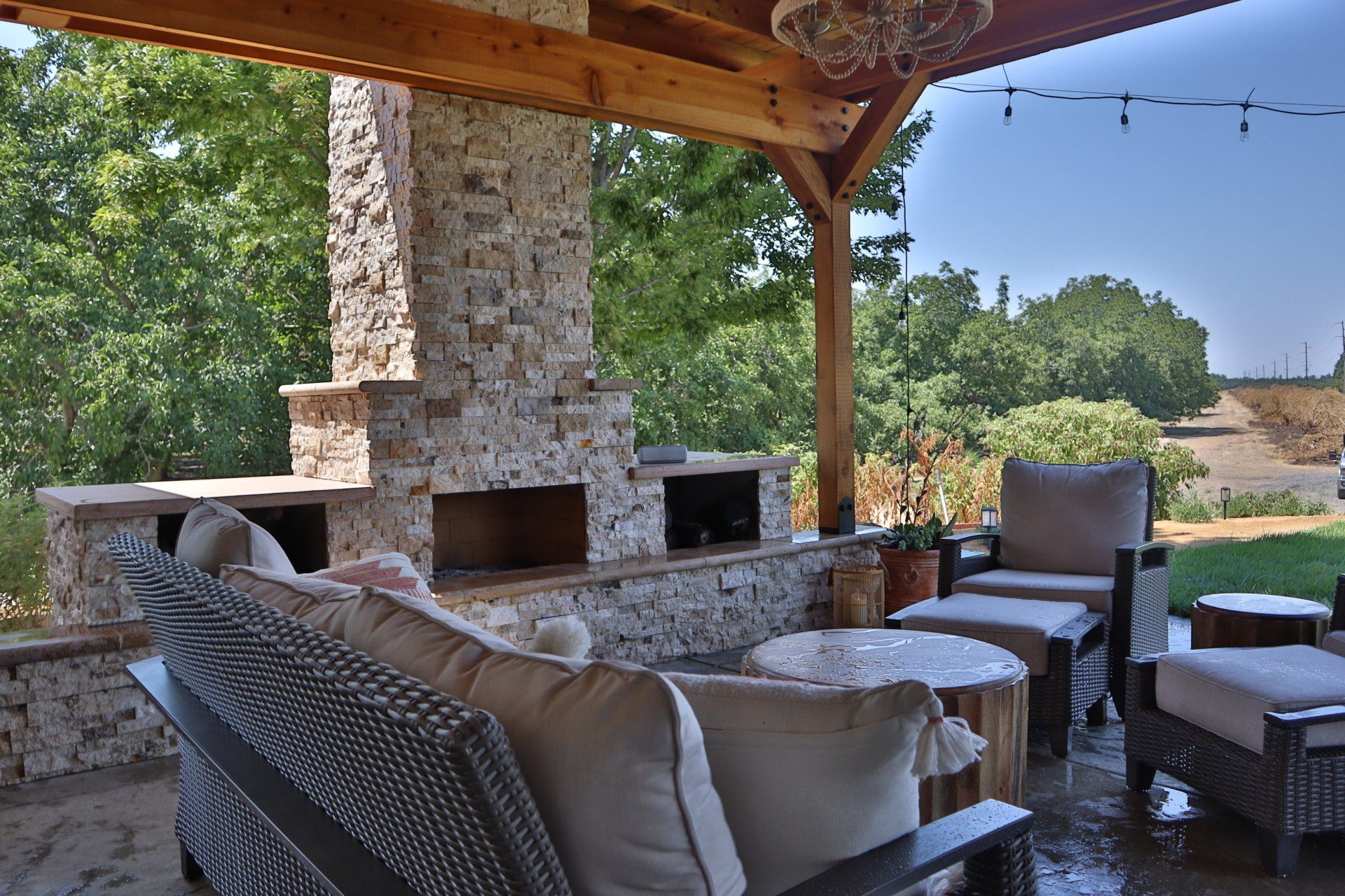 Patio with stone fireplace and seating area, wooden beams, and string lights, overlooking a green landscape.