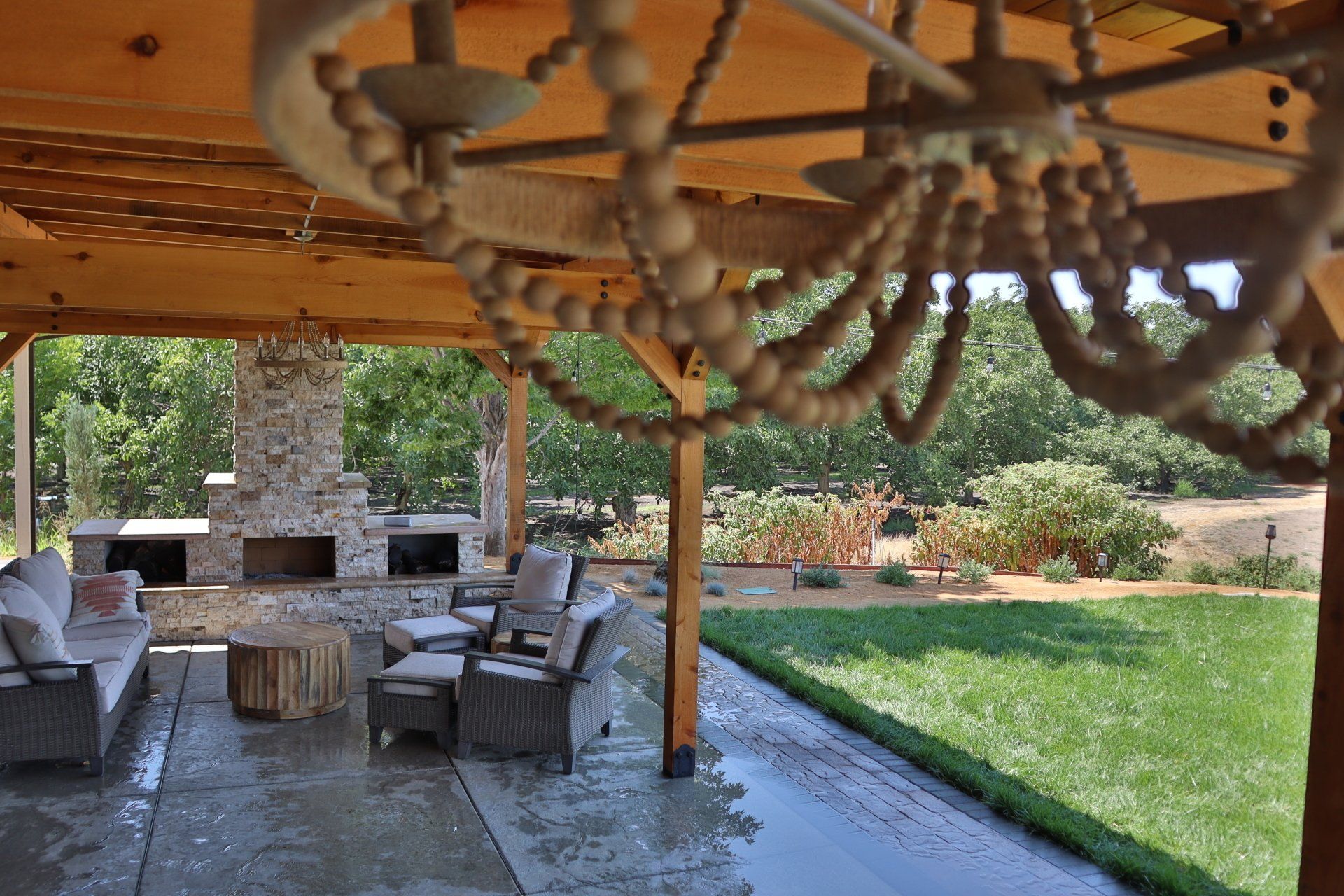 Covered patio with seating, fireplace, and chandelier overlooking a green lawn and trees.