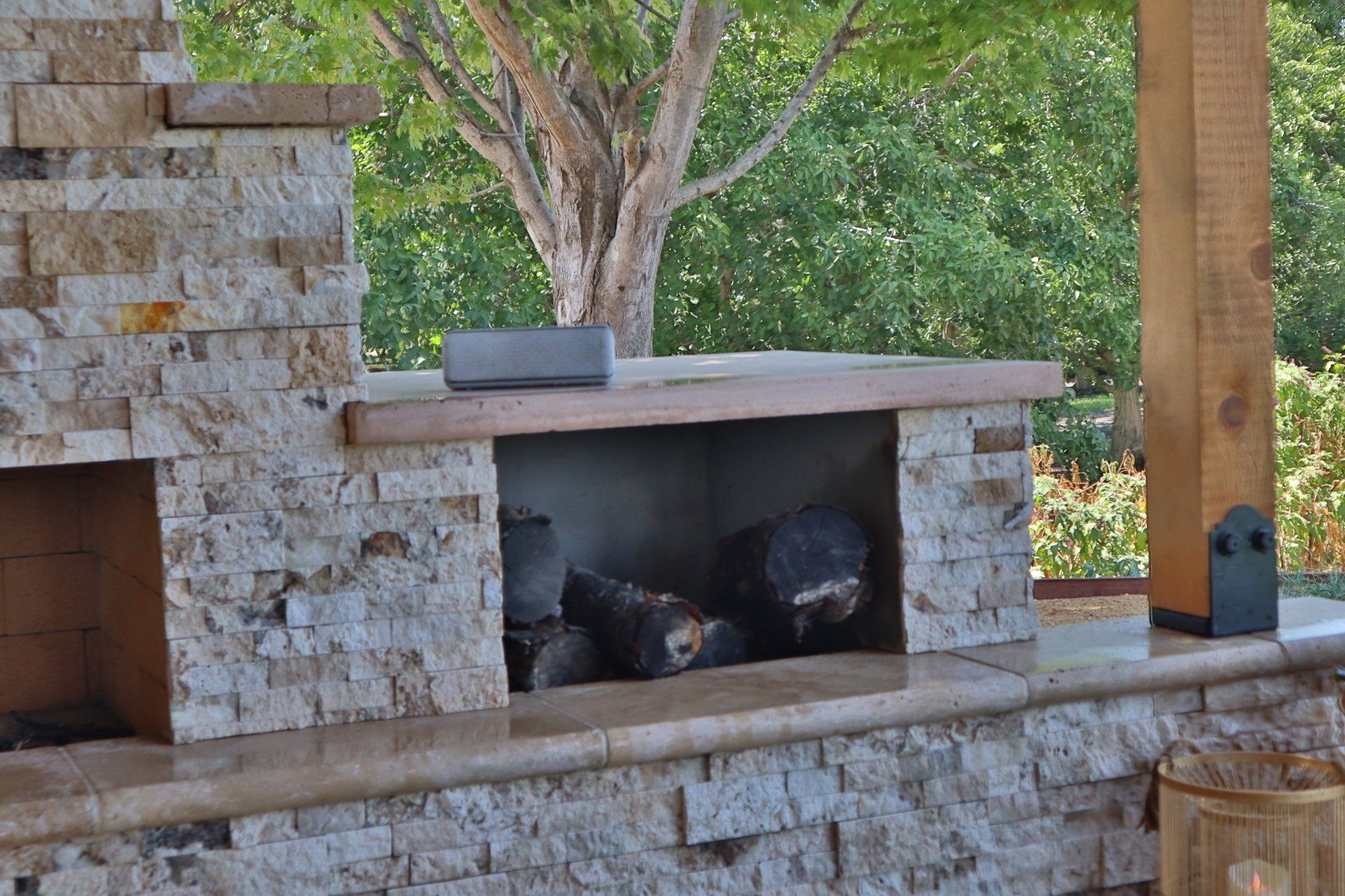 Stone outdoor fireplace with wood inside, next to a wooden post and greenery.