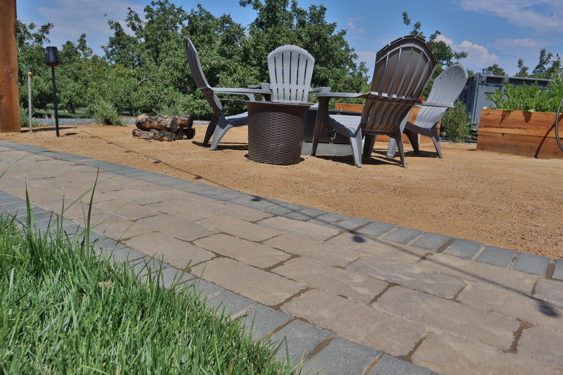 Patio with pavers and gravel, fire pit, Adirondack chairs, and garden beds under a blue sky.