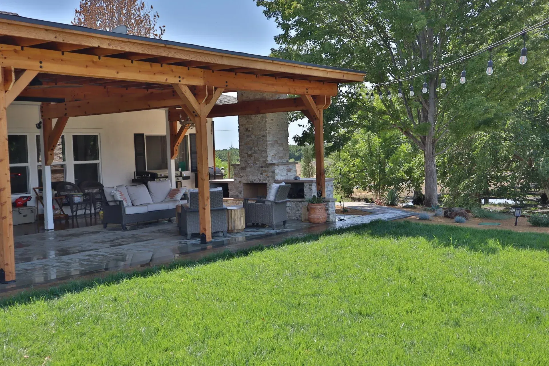 Covered patio with seating, fireplace, and green lawn. Wooden beams, neutral-colored house, and string lights are visible.