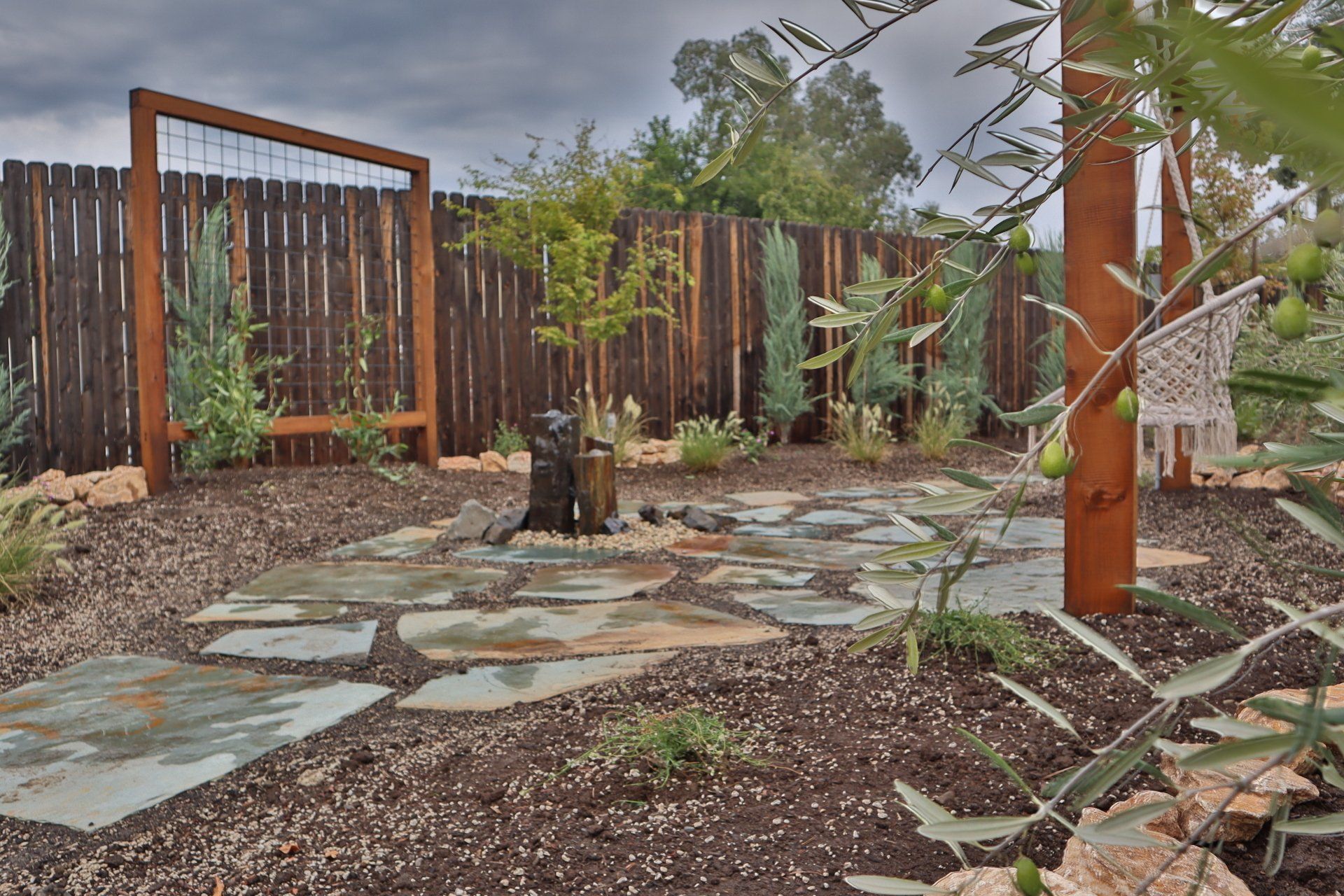 Stone patio in a garden with wooden fence, arbor, and olive tree. Brown and green tones.