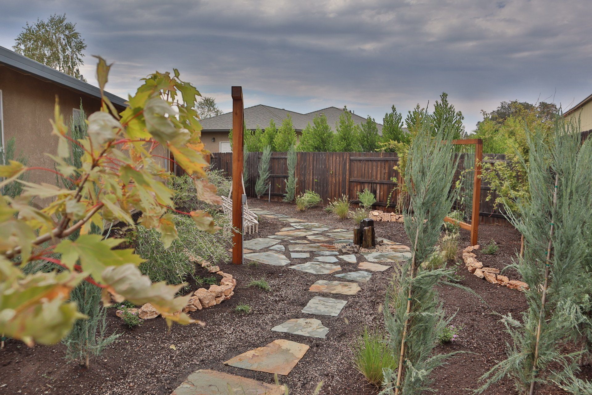 Stone pathway leads through a backyard garden with trees, plants, and a wooden gate under a cloudy sky.