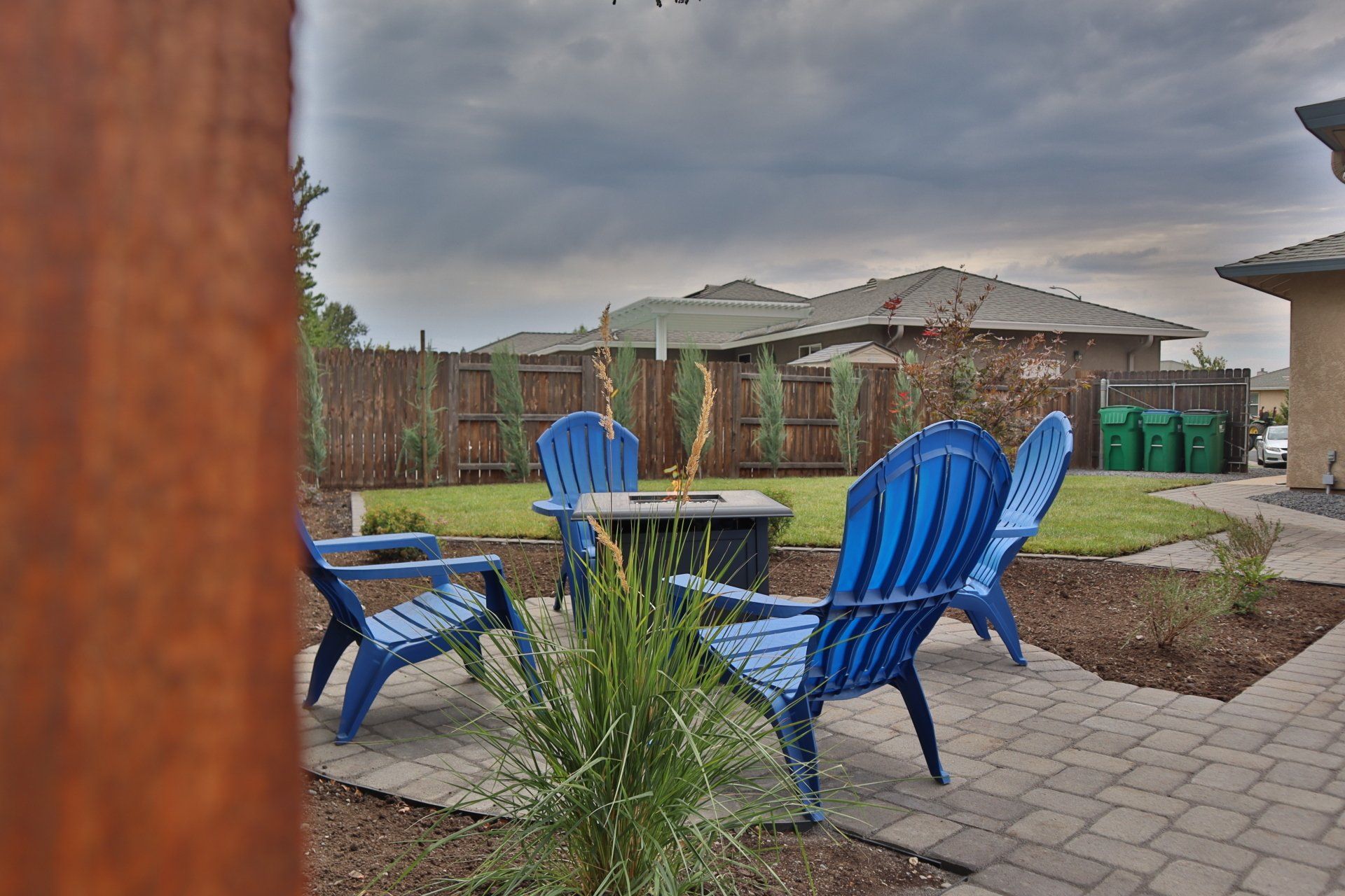 Backyard patio with blue chairs around a fire pit. Green grass, brown fence, and cloudy sky.