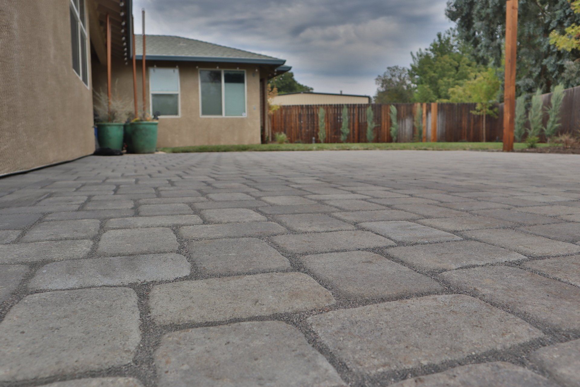 Gray brick patio in front of a tan house. A wooden fence and trees are visible in the background.