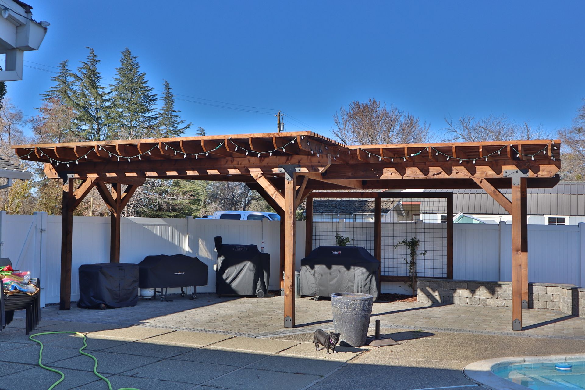 Wooden pergola over a patio with grills and a fence.