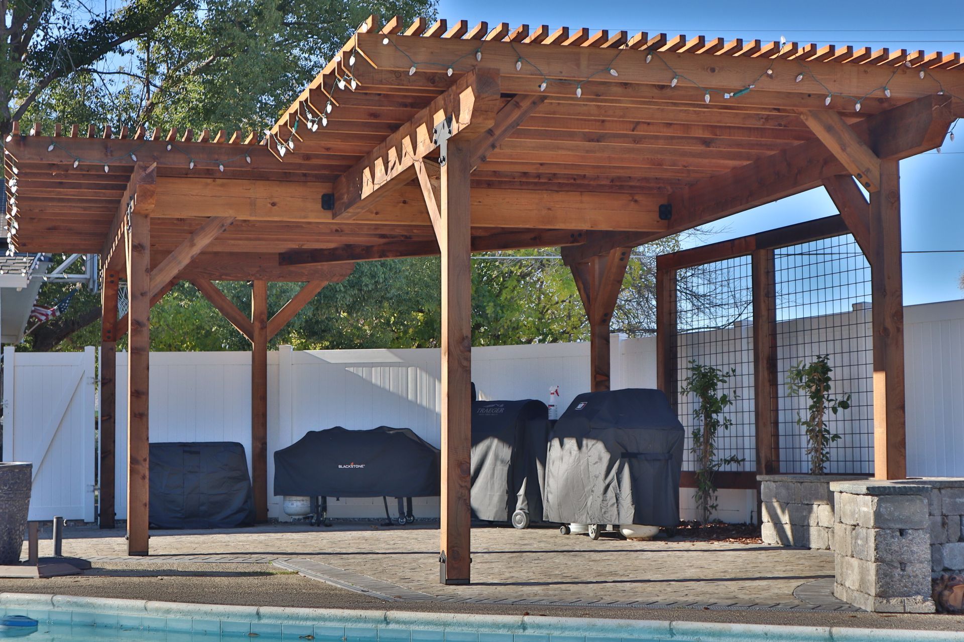 Wooden pergola over patio with grills, string lights, and a pool.