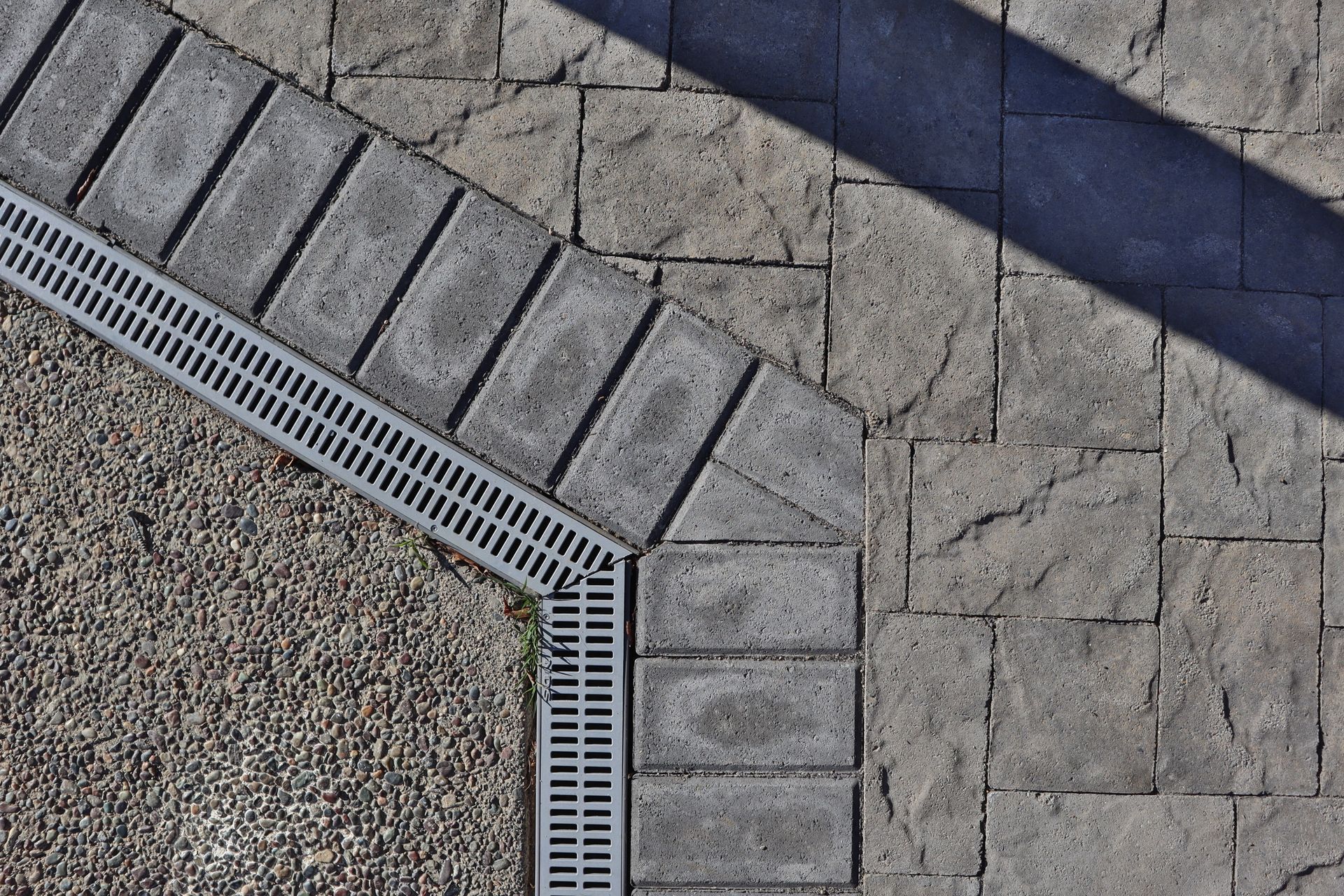 Overhead view of a sidewalk with a storm drain; paved with grey rectangular blocks and textured stone.