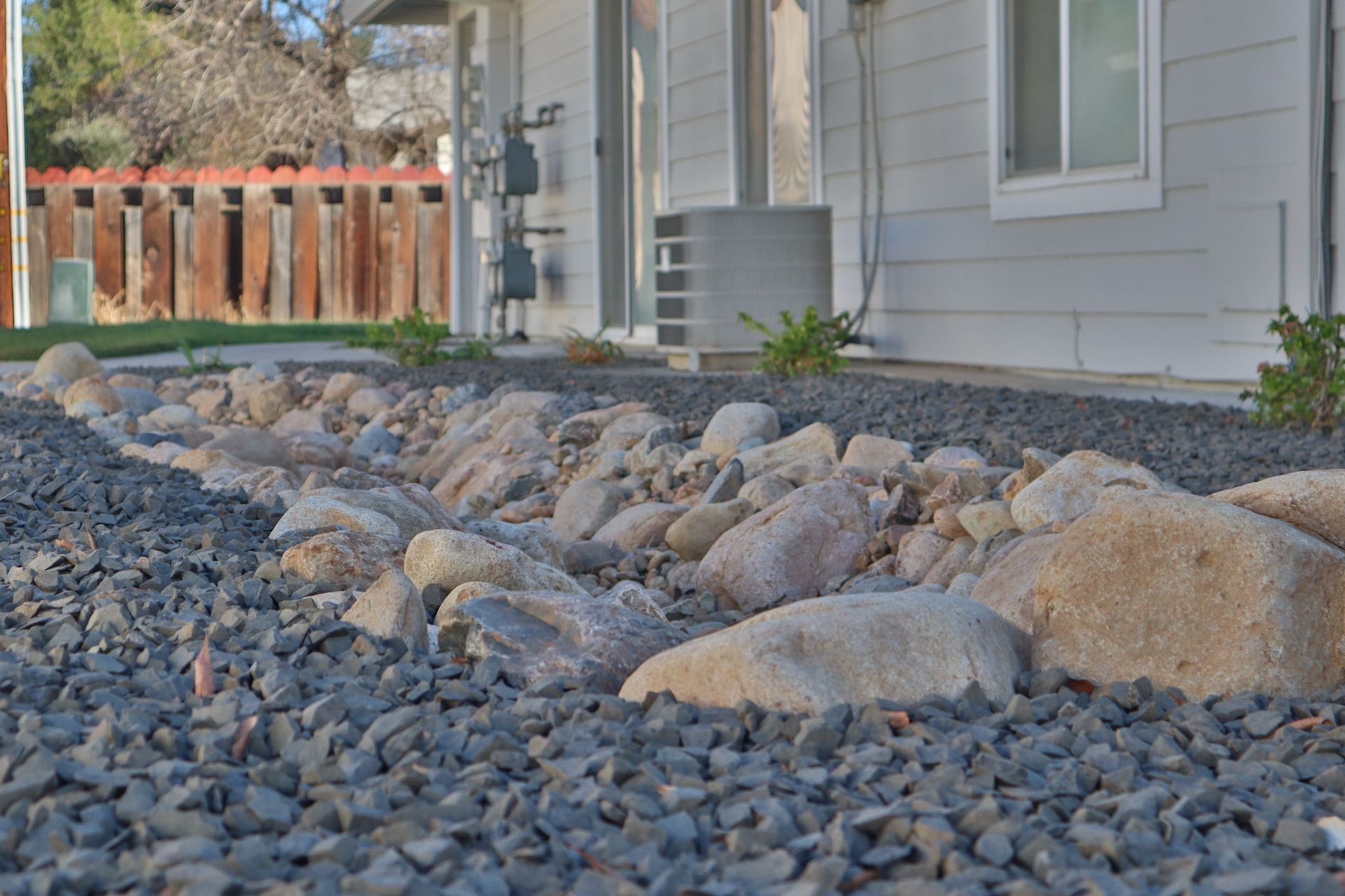 Rock border in front of a light-colored house. Gray gravel surrounds large, rounded rocks.