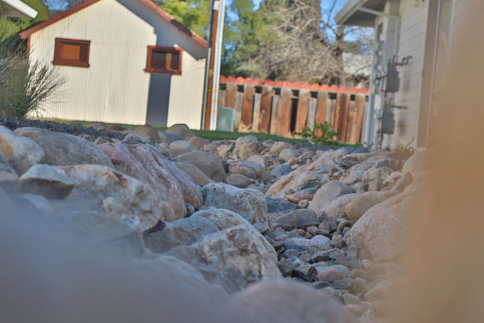 Rocks in foreground, small white building with brown trim, wooden fence in the background. Sunny day.