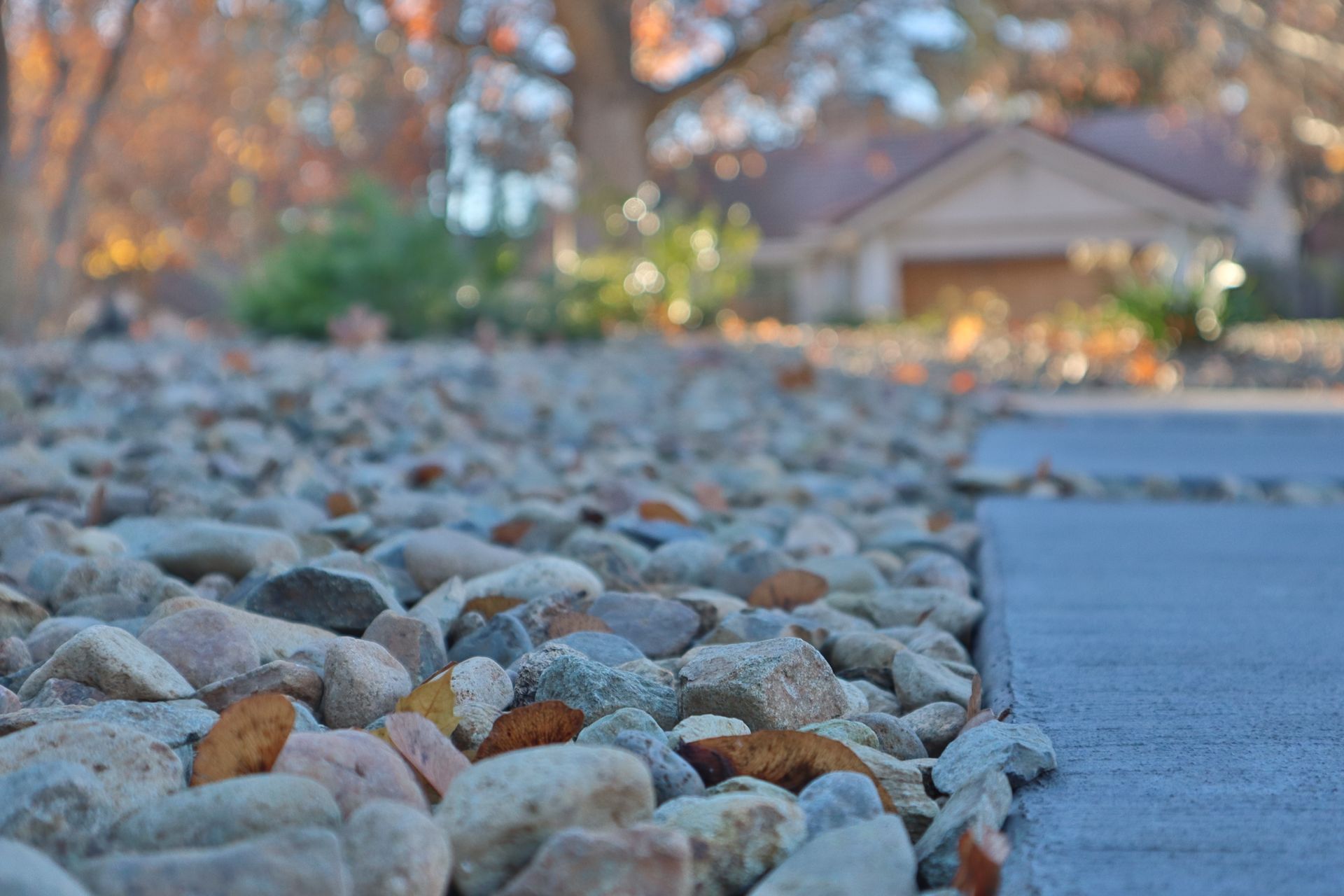 Gravel landscaping border next to a concrete walkway, with a house and tree in the blurred background.