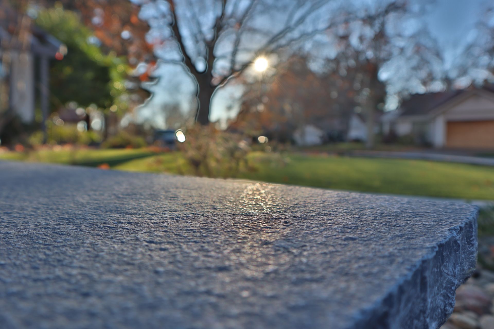 Close-up of a textured gray surface with sunlight reflecting. In the background, a residential area with houses and trees.