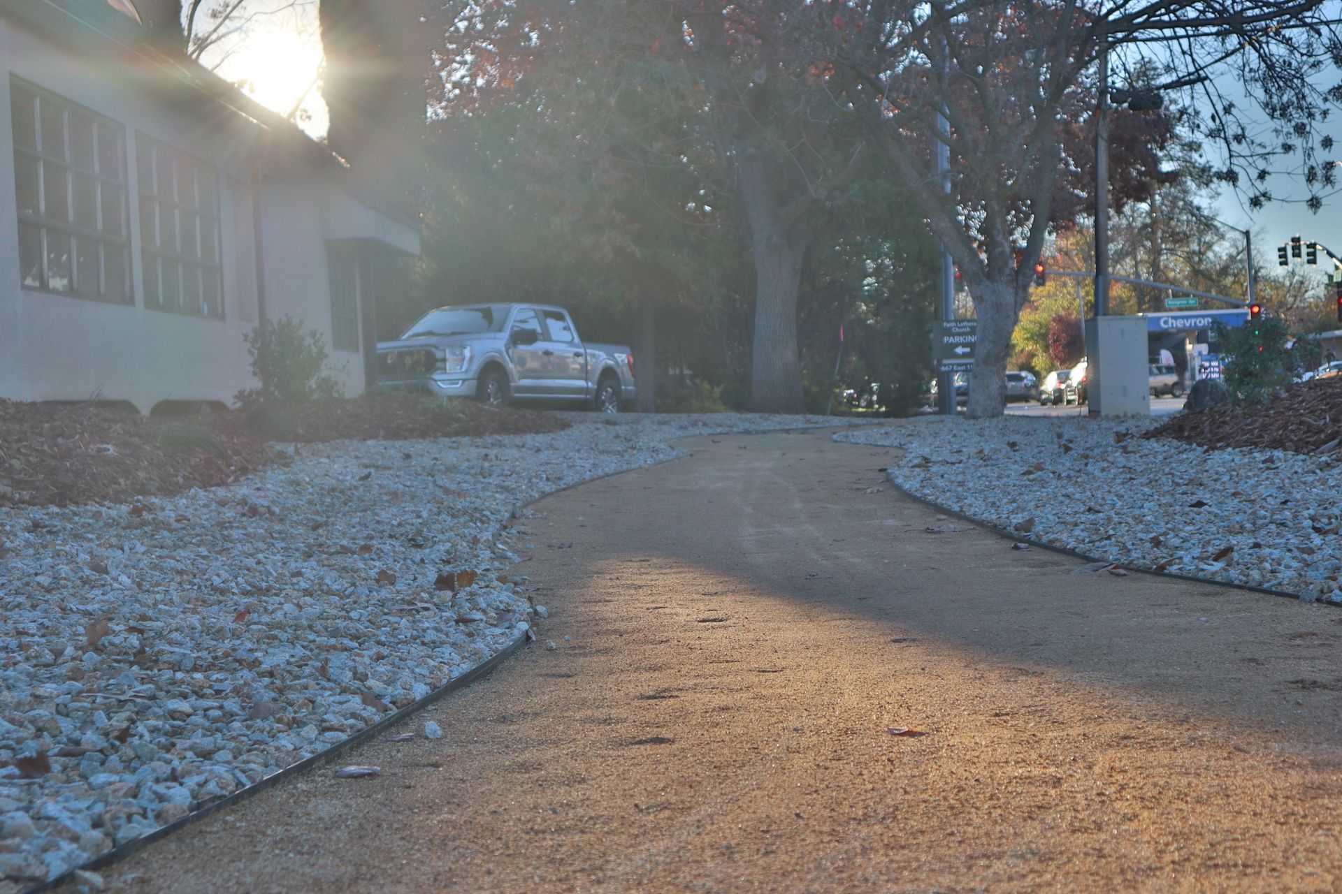 Gravel driveway curving towards building with parked truck, trees, and bright sunlight.