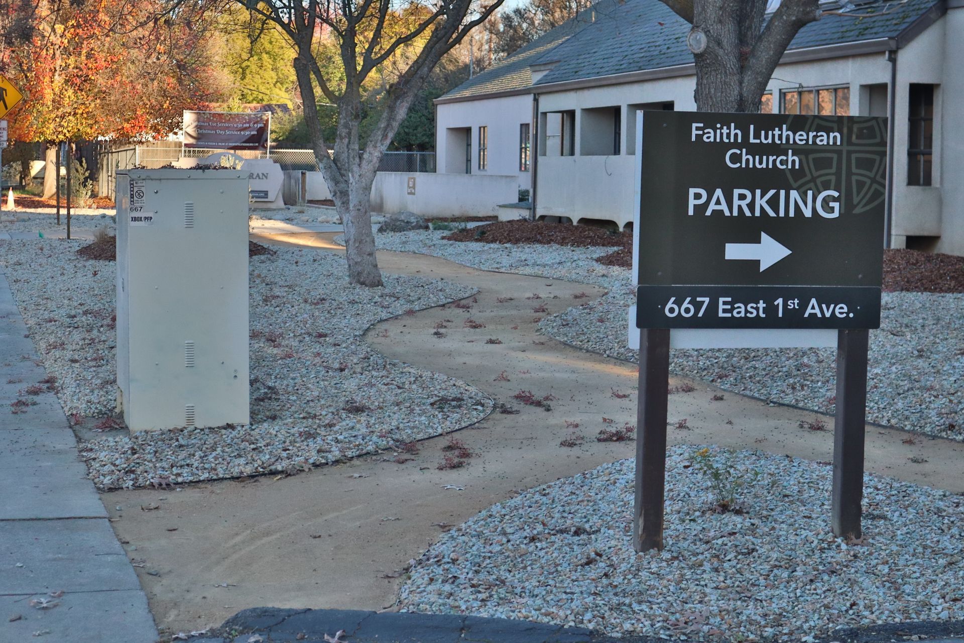 Sign for Faith Lutheran Church parking, arrow right. Gravel landscaping, beige building, street, trees.