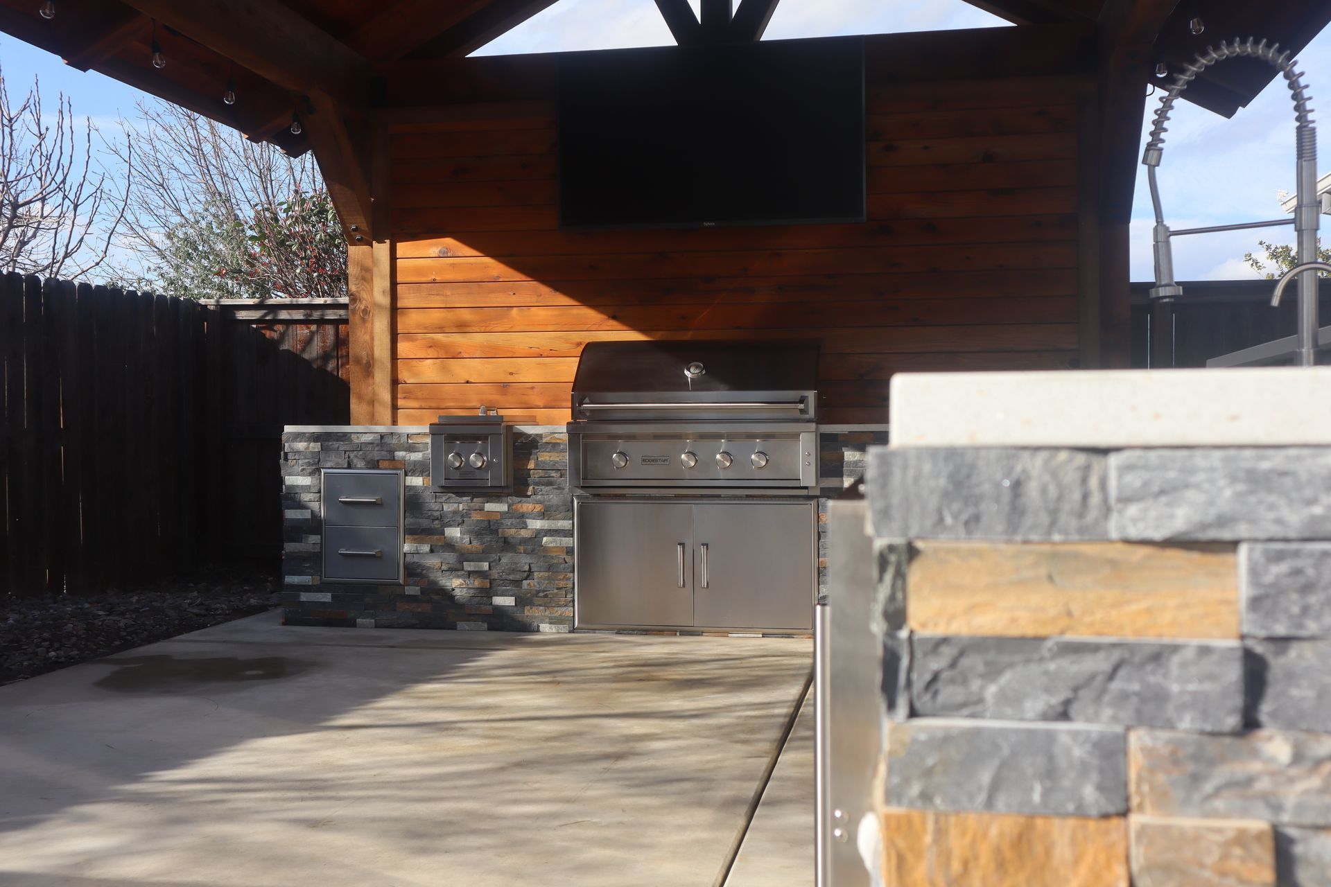 Outdoor kitchen with grill, stone accents, and TV mounted under a wooden pergola.