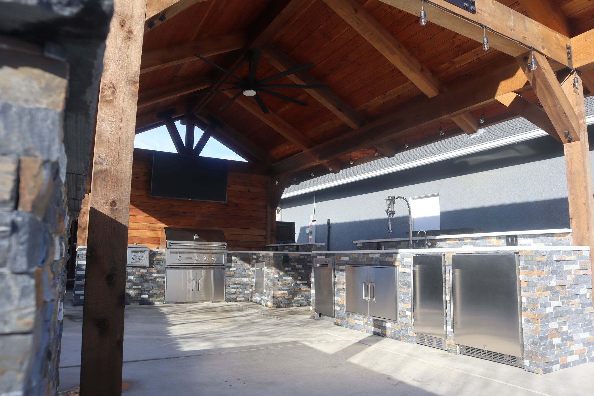 Outdoor kitchen with wooden roof, stone accents, stainless steel appliances, and a concrete patio.